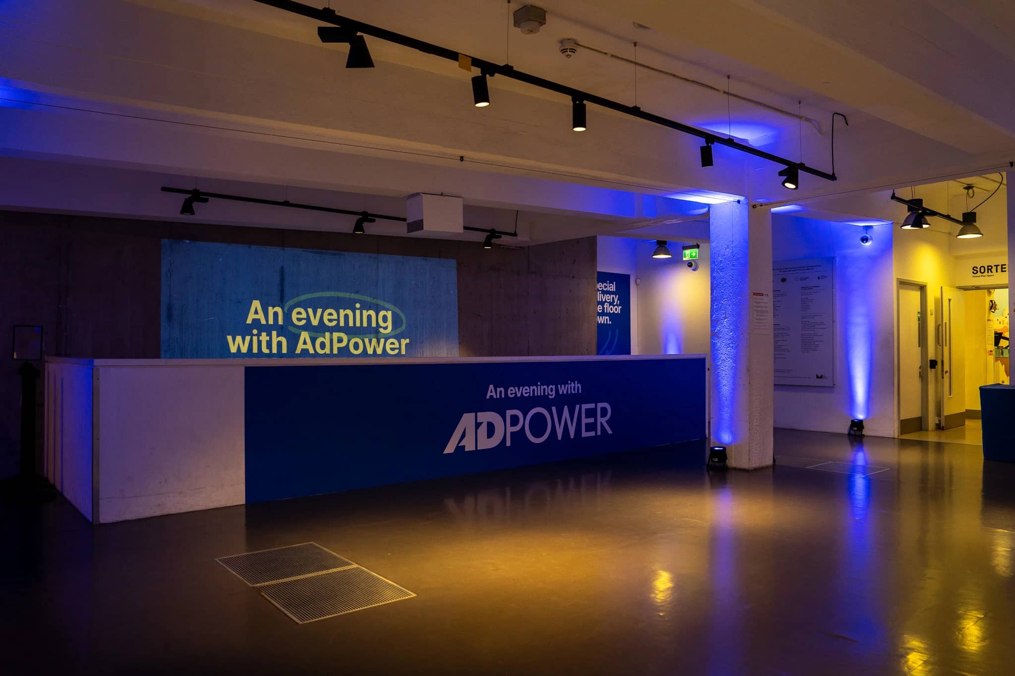 AdPower branding across the low wall in the welcome space, with blue uplighting and the event title projected behind.