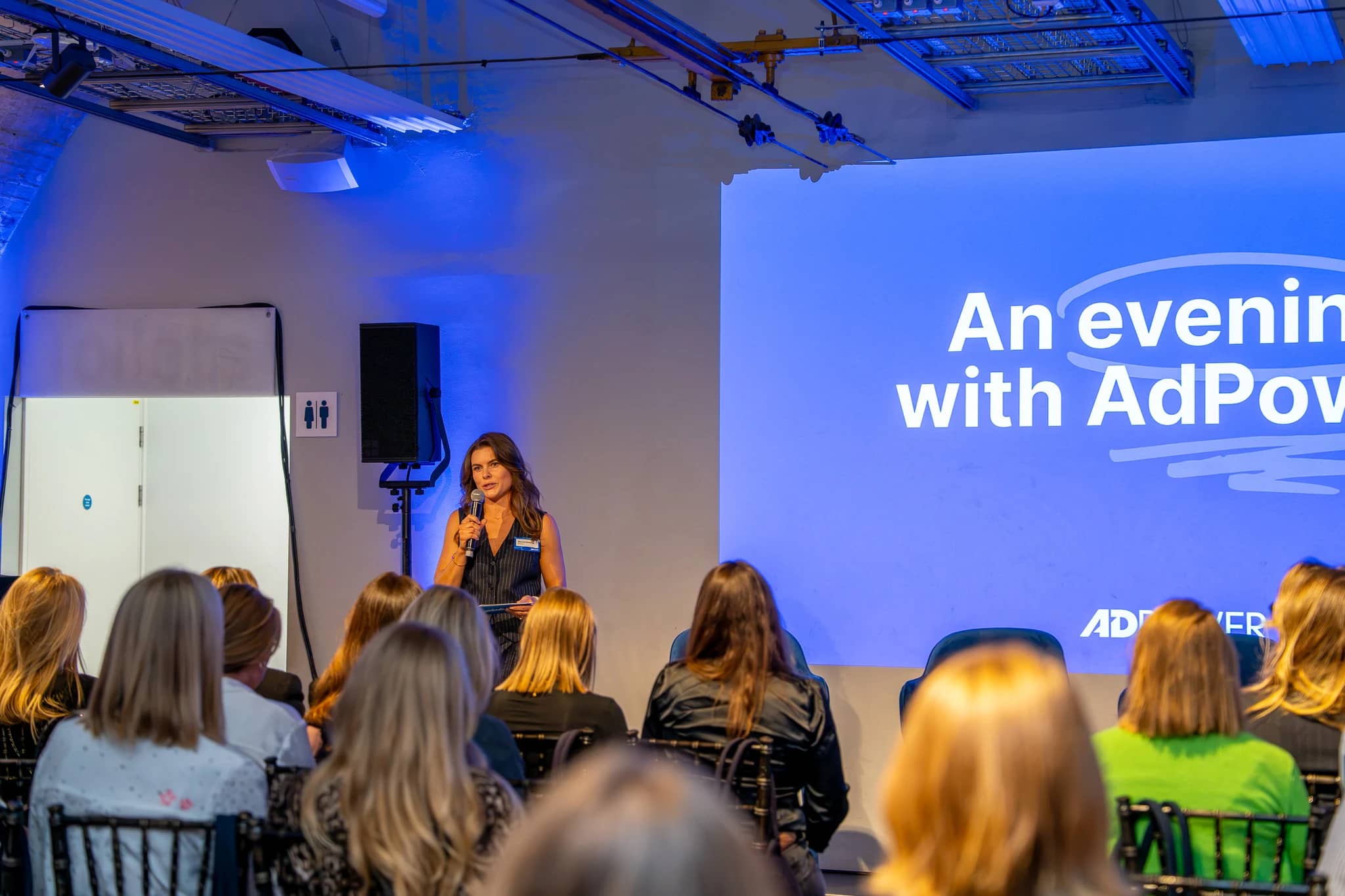 A speaker addresses the seated AdPower audience in front of the event projection and blue-lit brickwork at Mail Rail.