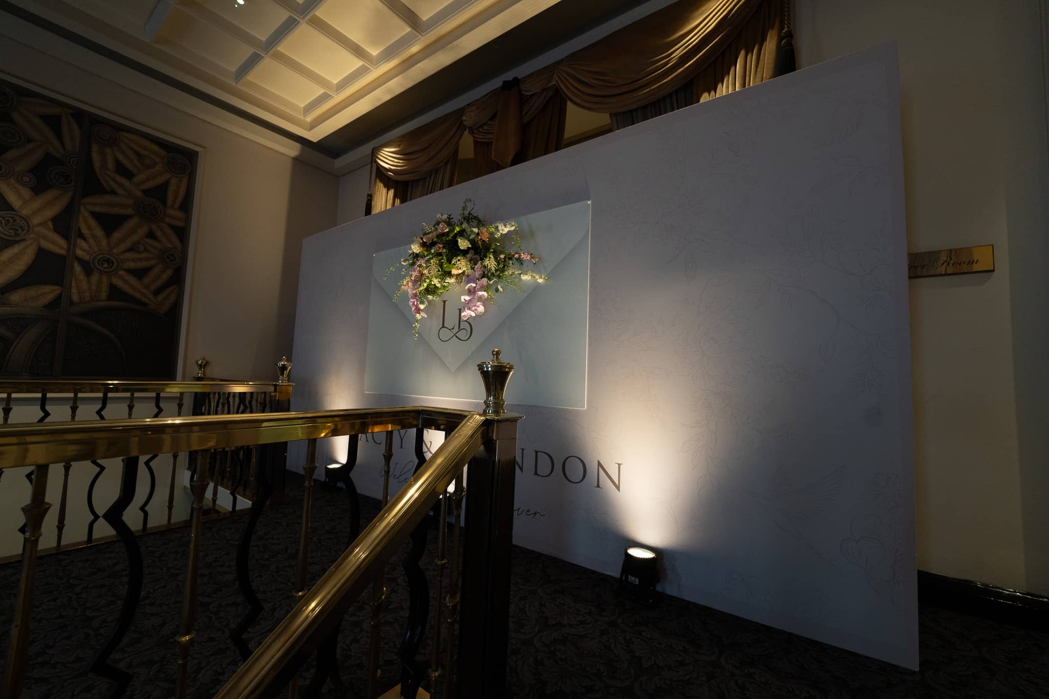 The wedding media wall seen from the staircase at The Savoy, floral spray mounted on the envelope motif and lit by a floor uplighter, by Kudos AV