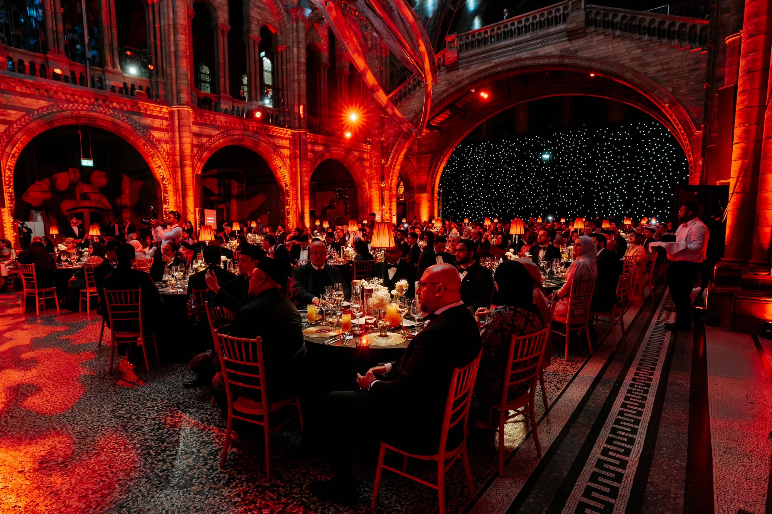 Tables set for the gala dinner in Hintze Hall under red uplighting on the arches, Awakening Music 25th anniversary at the Natural History Museum, lit by Kudos AV.