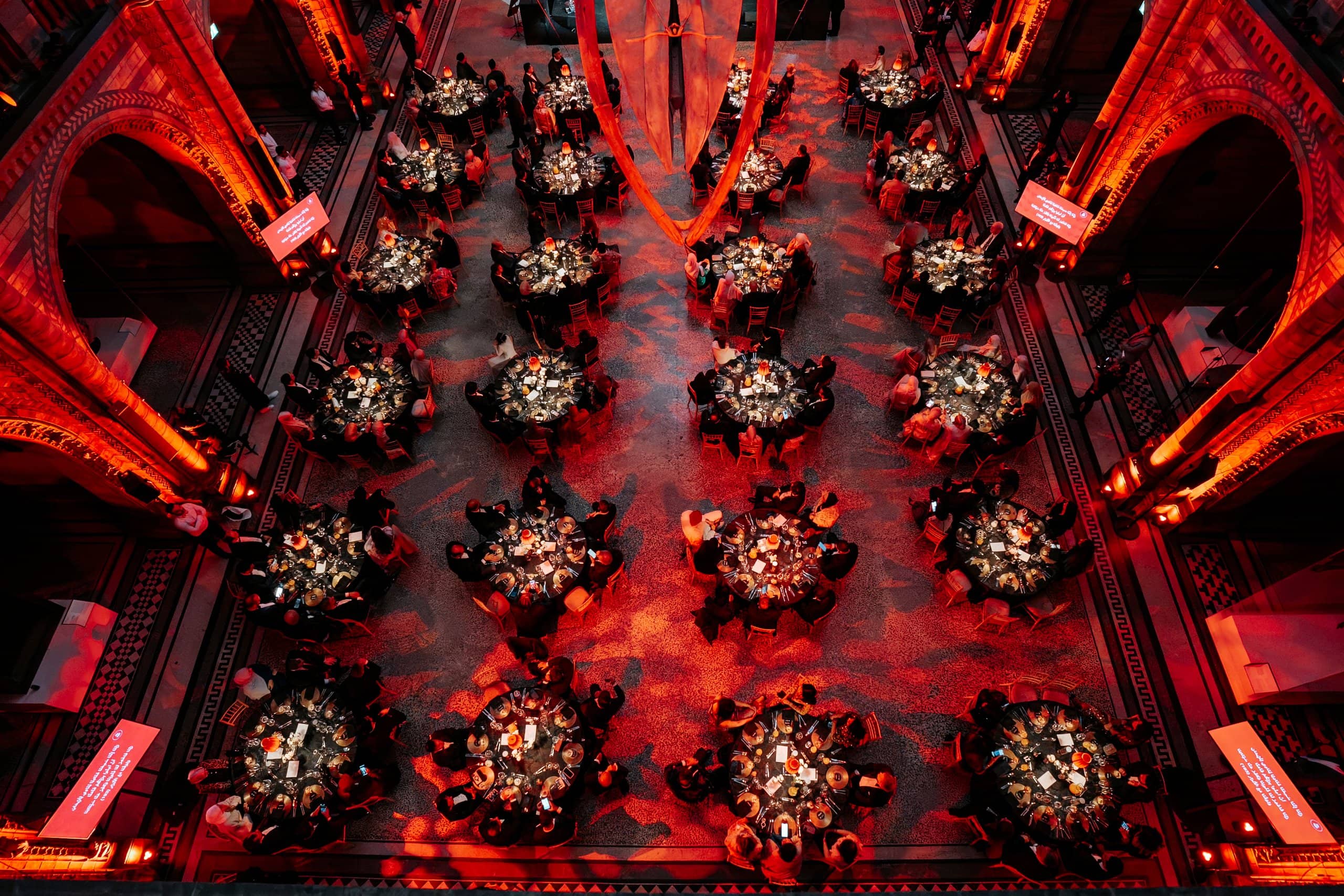 Hintze Hall from above, round tables with florals set for the Awakening Music 25th anniversary dinner under red uplighting, by Kudos AV.