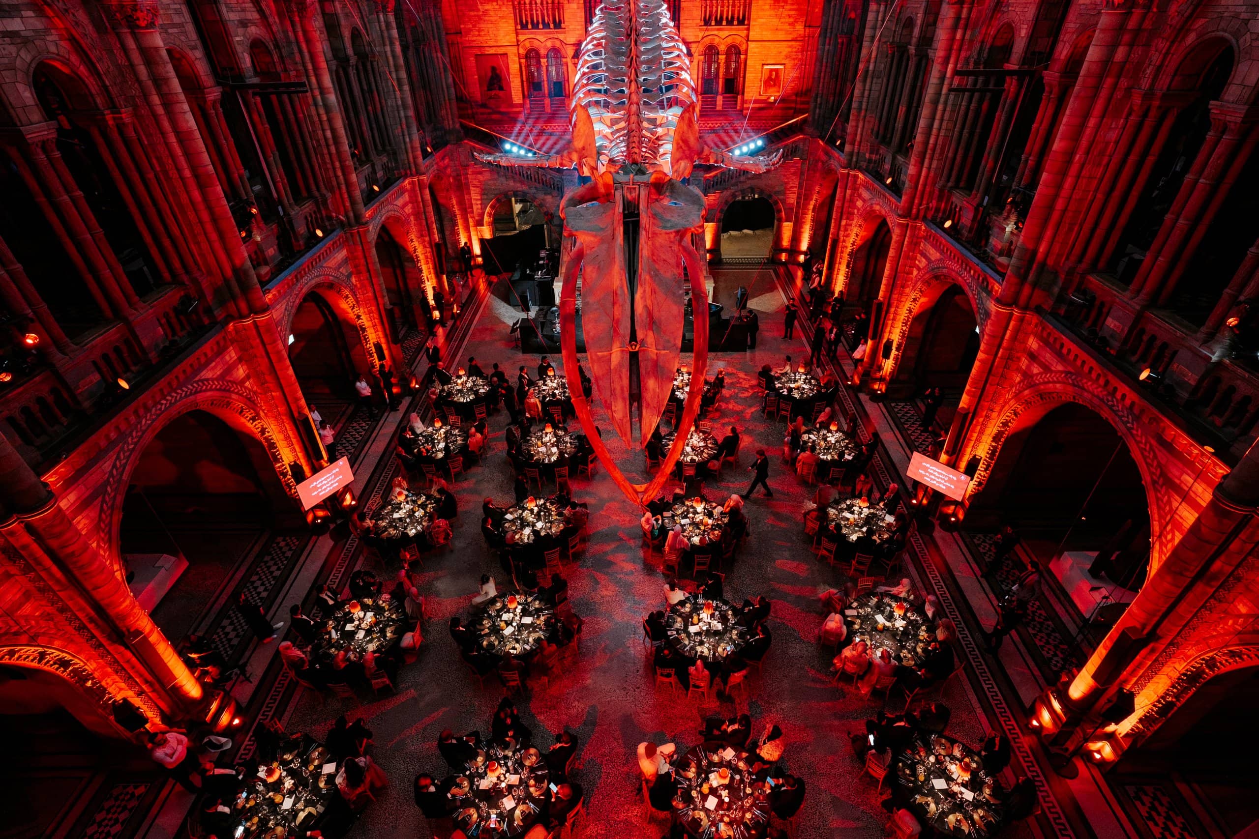 Looking down on Hintze Hall, Hope the blue whale above round tables lit red for the Awakening Music 25th anniversary, by Kudos AV.