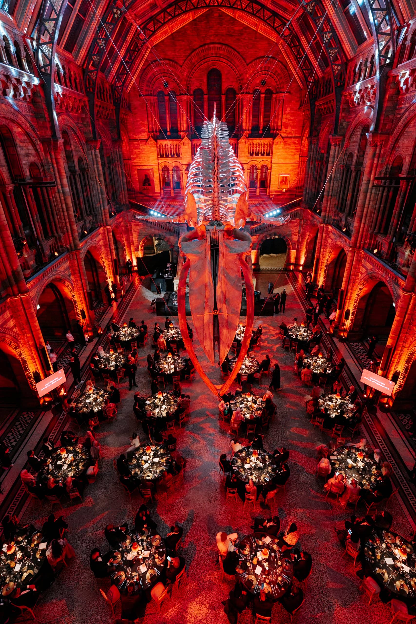 Hintze Hall lit red from floor to vault with dinner tables set through the hall for the Awakening Music 25th anniversary at the Natural History Museum, by Kudos AV.