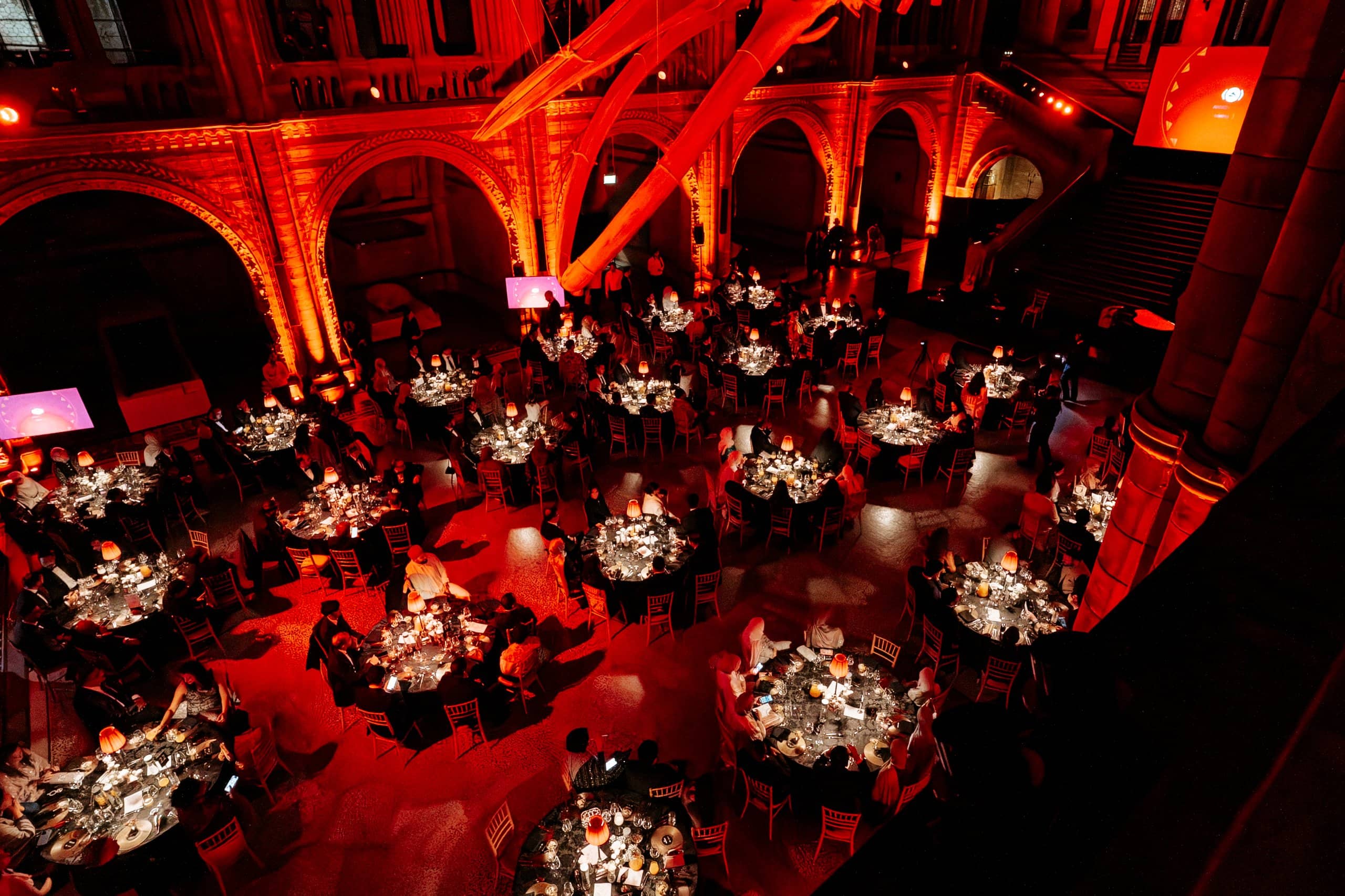 Hintze Hall from above, dressed dinner tables and the arches lit red for the Awakening Music 25th anniversary, by Kudos AV.