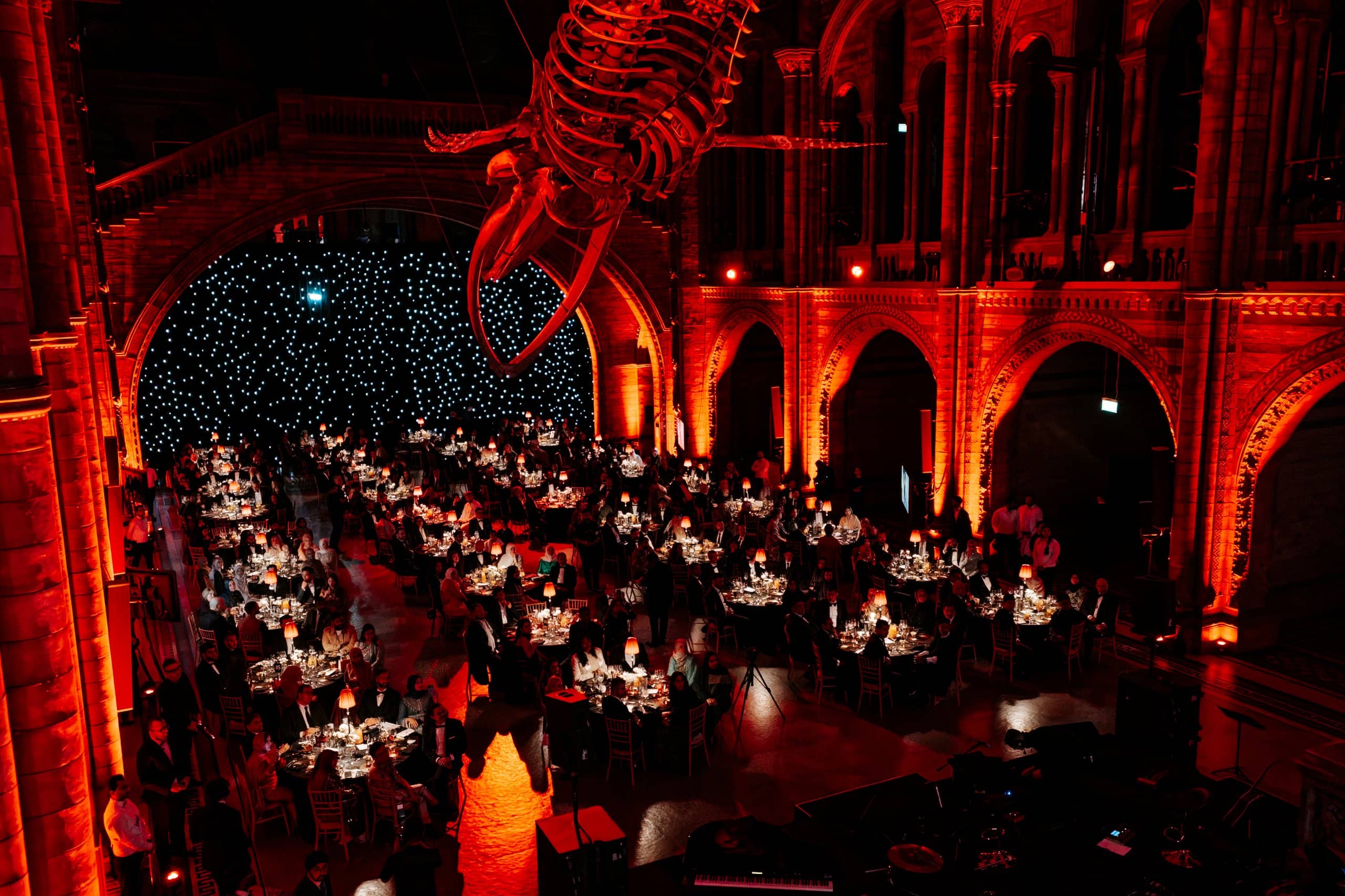 Round tables under red uplighting with Hope the blue whale overhead in Hintze Hall, Awakening Music 25th anniversary dinner by Kudos AV.