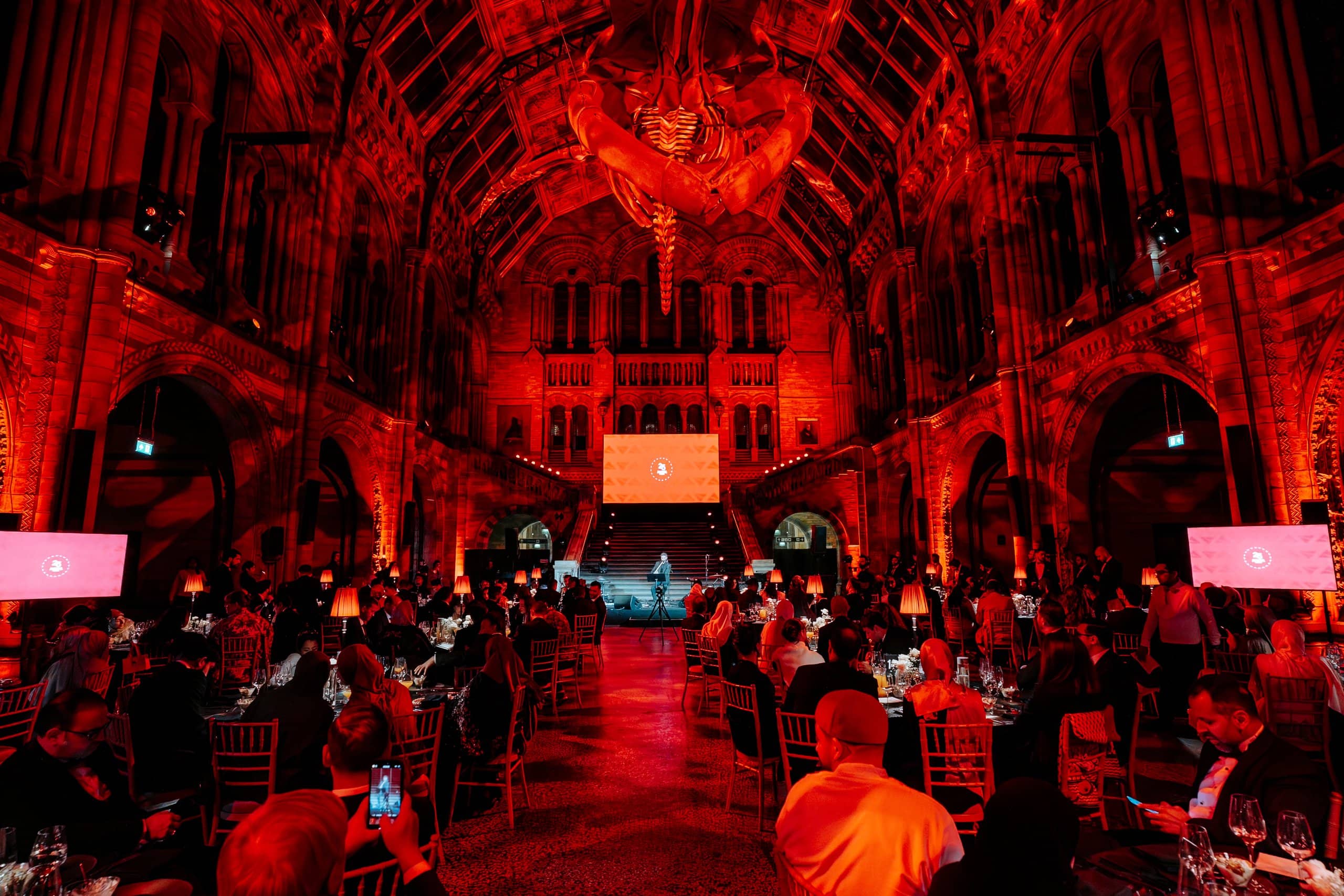 Hintze Hall set for the Awakening Music 25th anniversary: red uplighting, dinner tables, and the stage and LED screen by Kudos AV.