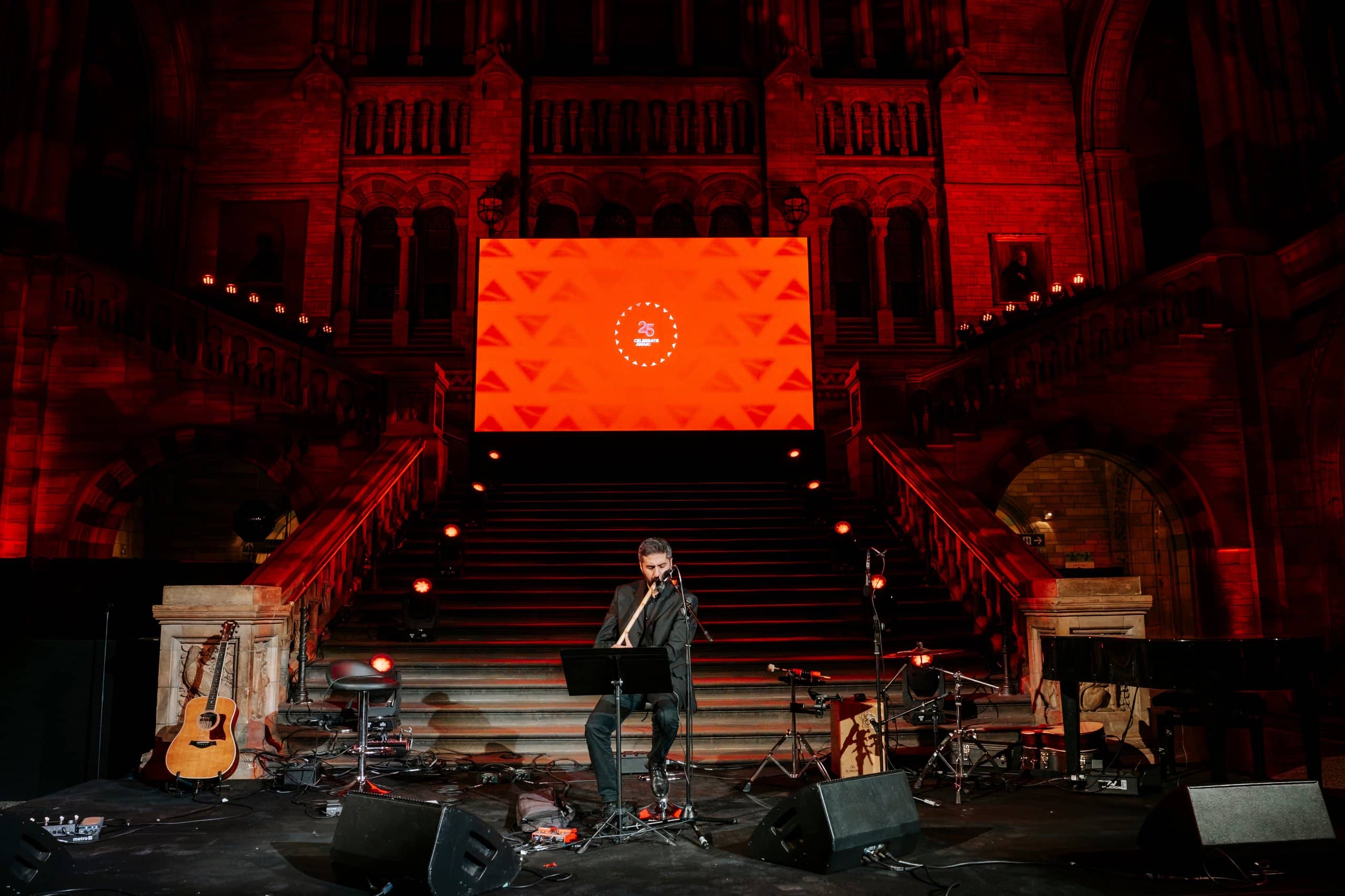 A performer on stage with acoustic guitars in front of the LED screen, red uplighting on the Hintze Hall arches, Awakening Music 25th anniversary by Kudos AV.