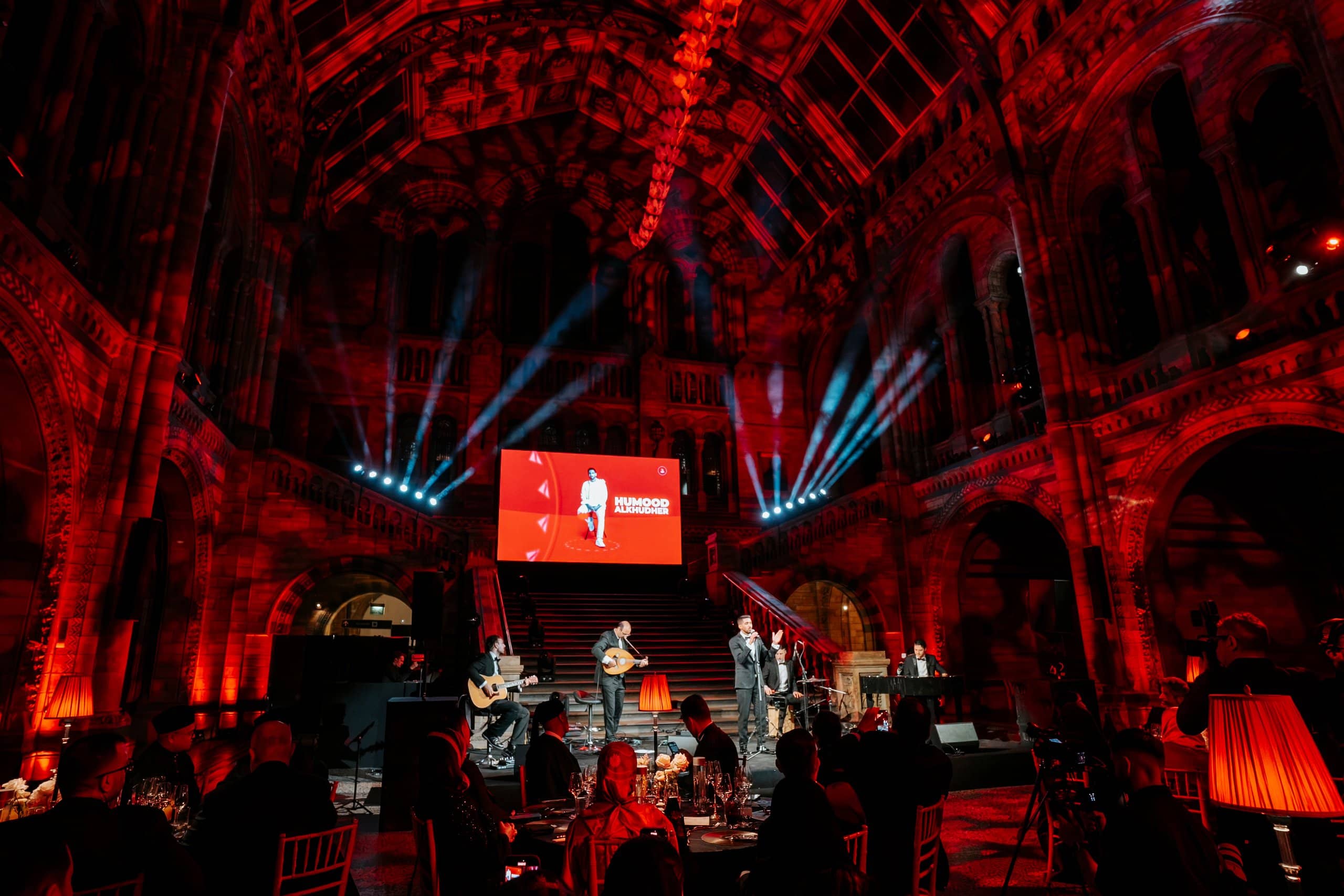The band performing on stage with blue beams and red uplighting in Hintze Hall, the LED screen behind, Awakening Music 25th anniversary by Kudos AV.