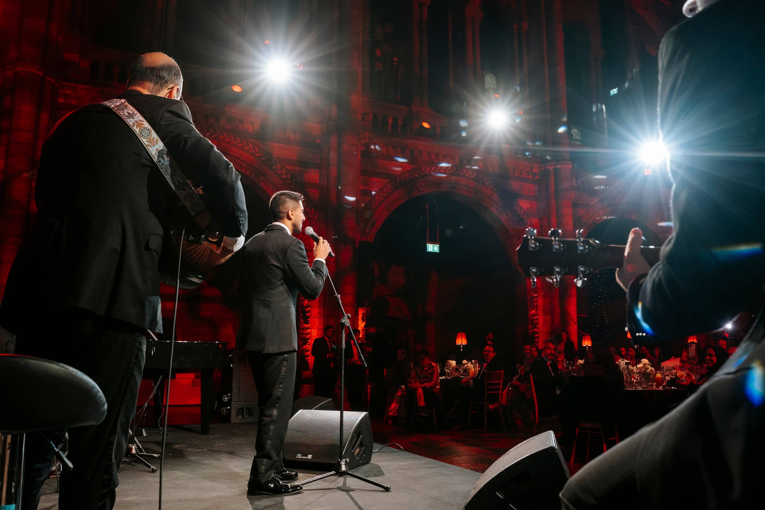 Two speakers at the microphone on stage under spotlights and red uplighting, Awakening Music 25th anniversary in Hintze Hall, by Kudos AV.