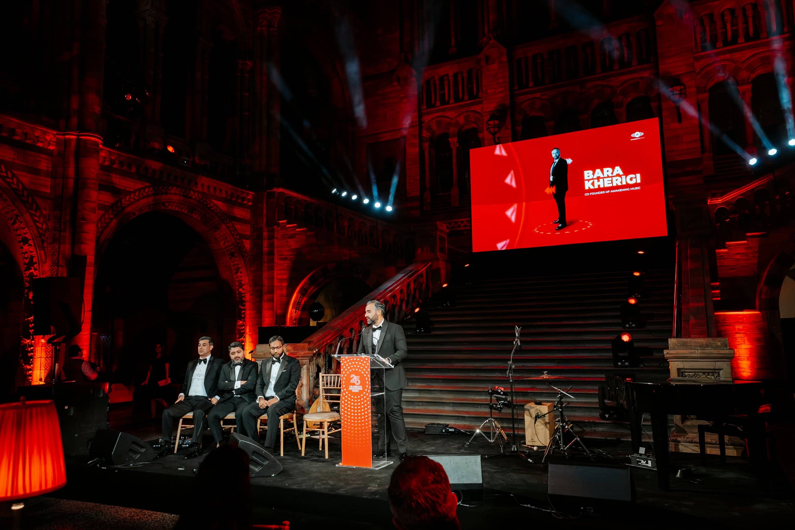 A speaker at the lectern with three panellists seated on stage and the LED screen above, Awakening Music 25th anniversary in Hintze Hall by Kudos AV.