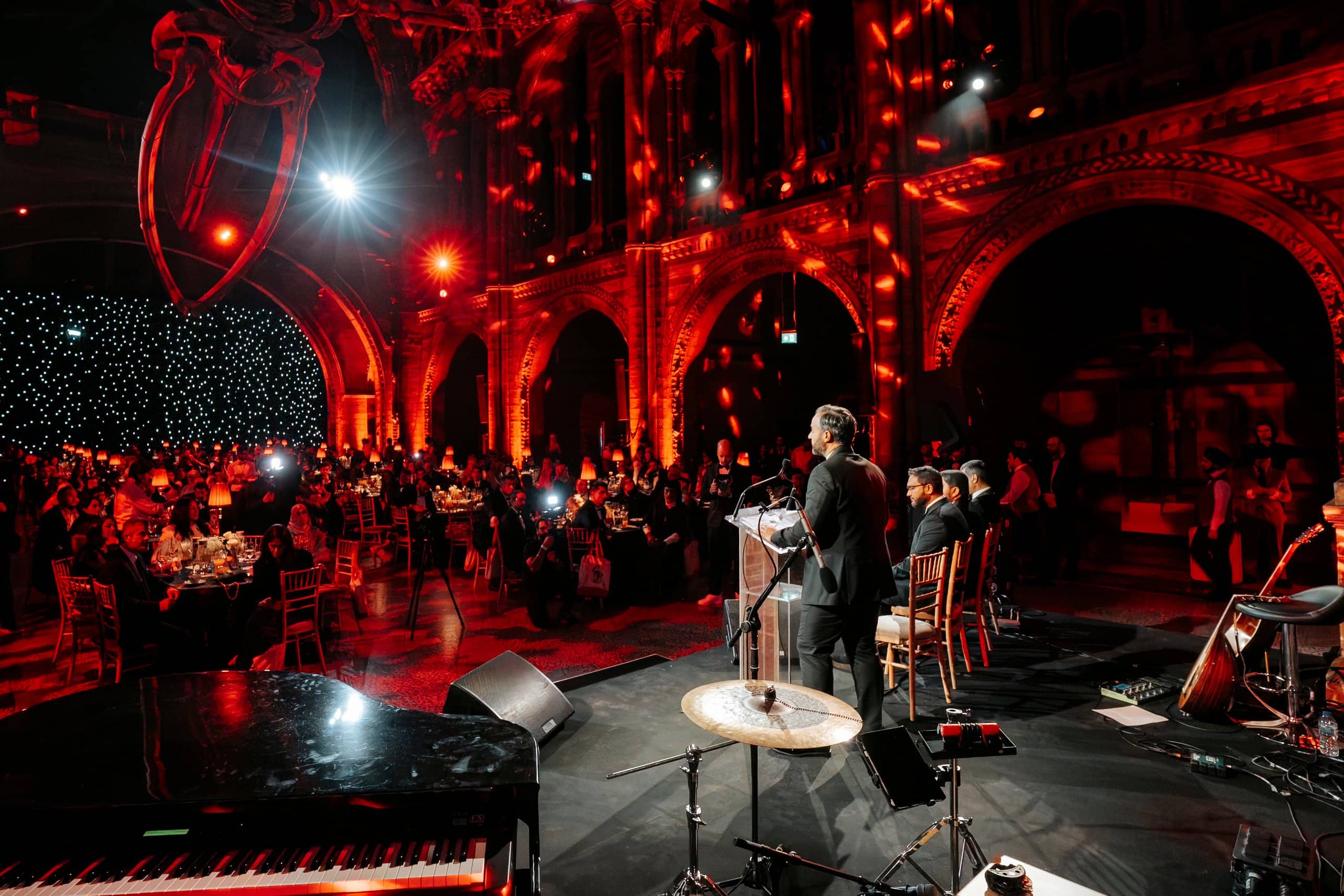 A grand piano on stage and guests at candlelit tables under red uplighting on the Hintze Hall arches, Awakening Music 25th anniversary by Kudos AV.