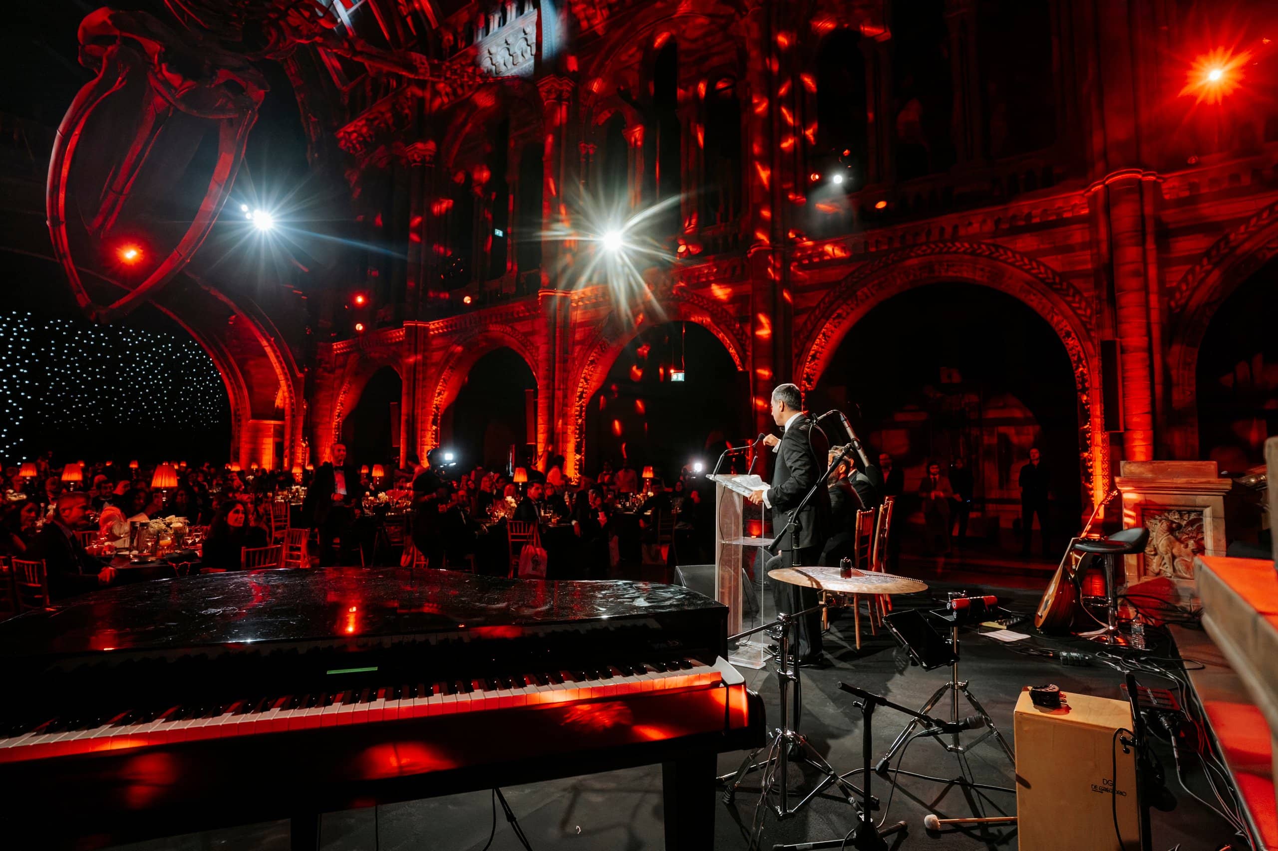 A speaker at the lectern beneath the red lit stone arches of Hintze Hall, guests seated at dinner, Awakening Music 25th anniversary by Kudos AV.