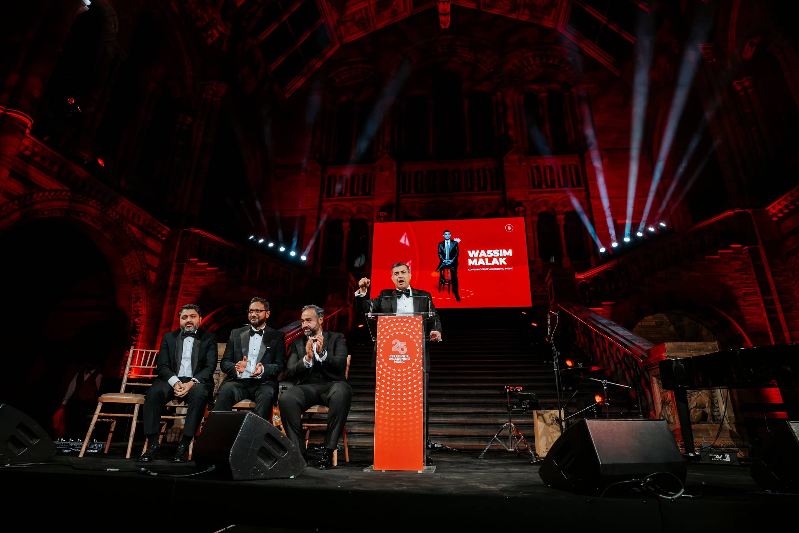Four guests on stage for a presentation, the LED screen and red uplighting behind, Awakening Music 25th anniversary in Hintze Hall by Kudos AV.