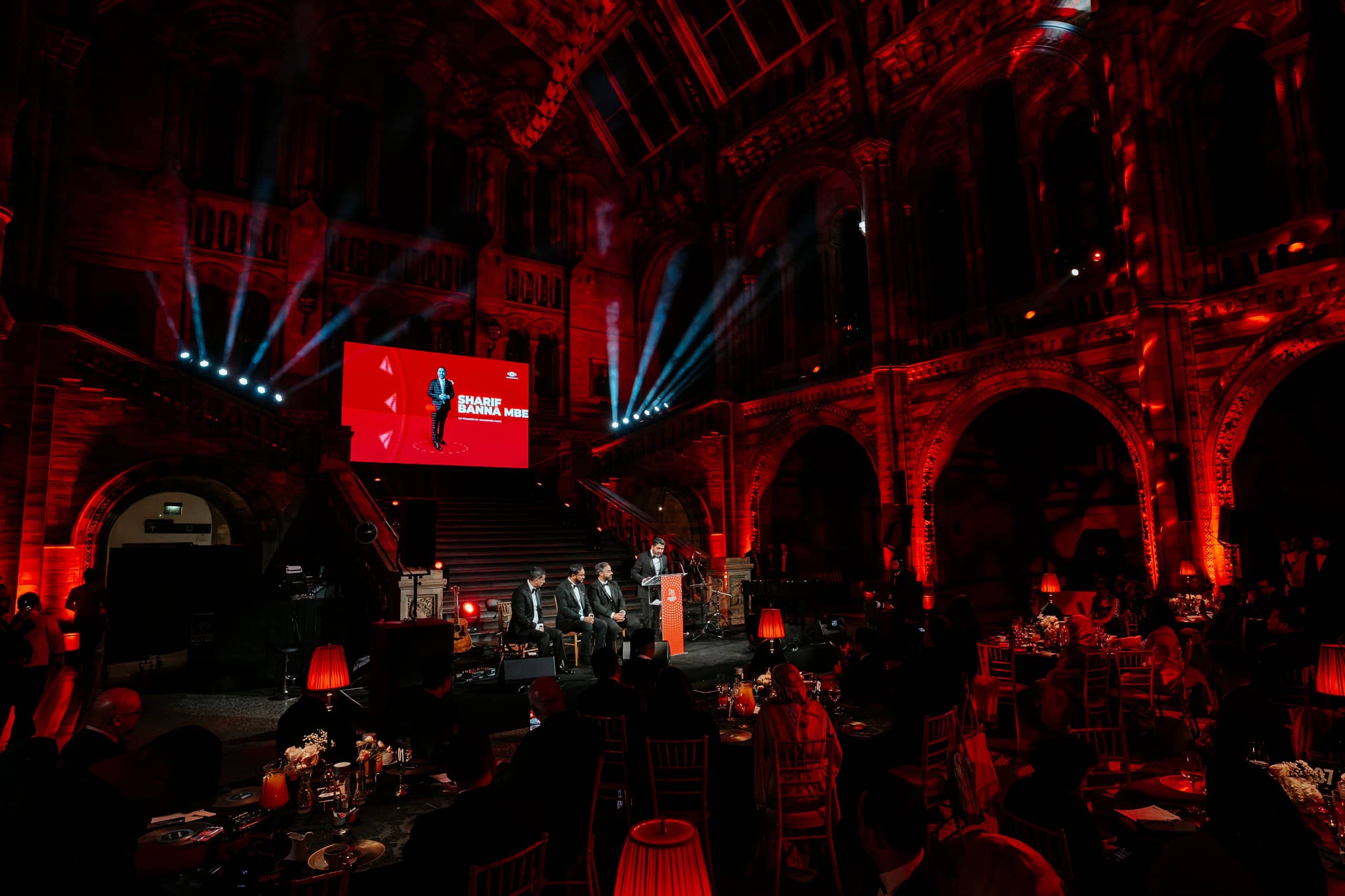 The stage and LED screen framed by red uplighting across Hintze Hall during the Awakening Music 25th anniversary dinner, by Kudos AV.
