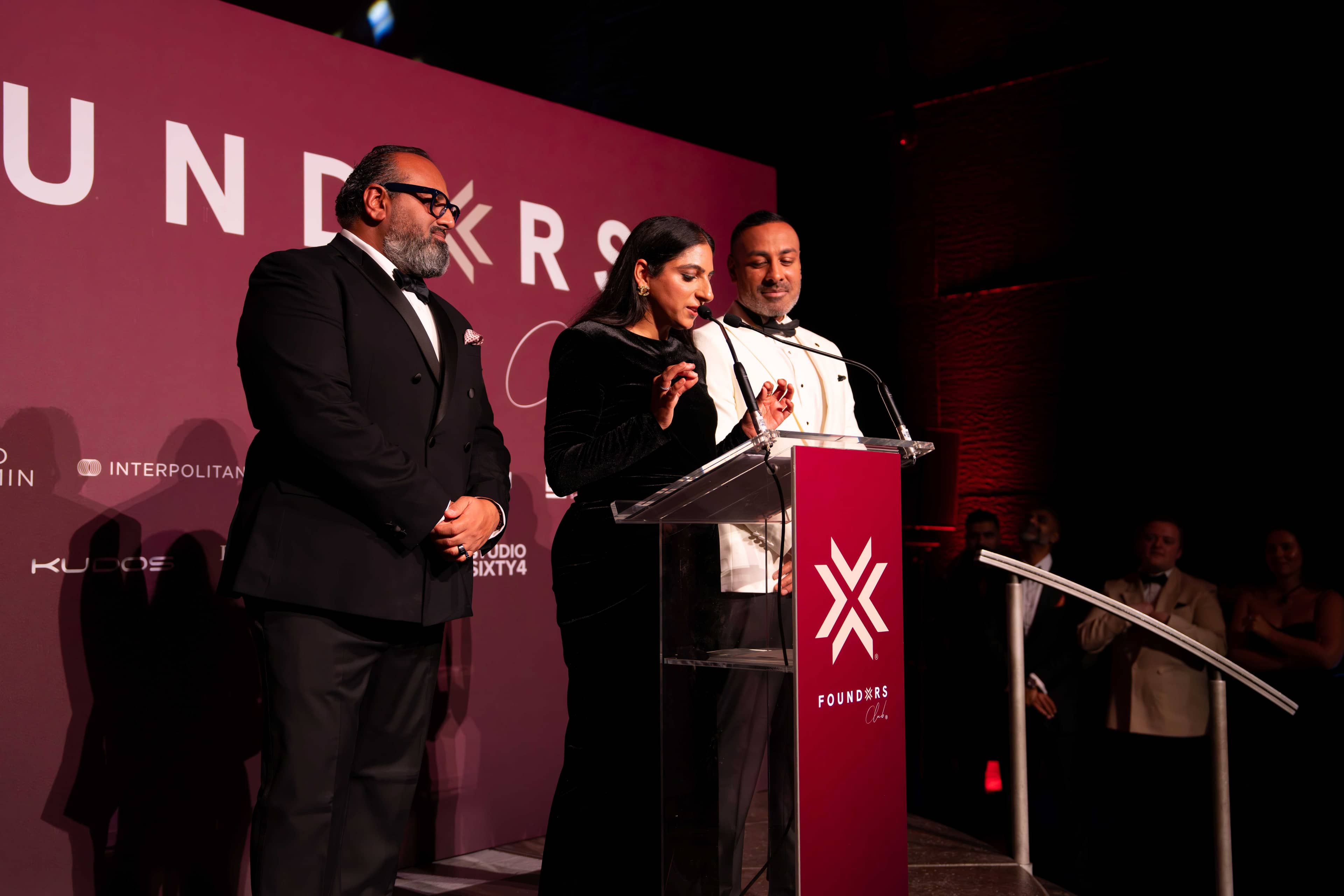 Three people at the red FOUNDXRS lectern on stage under the stage wash, FOUNDXRS Club black tie party in Earth Hall, by Kudos AV.
