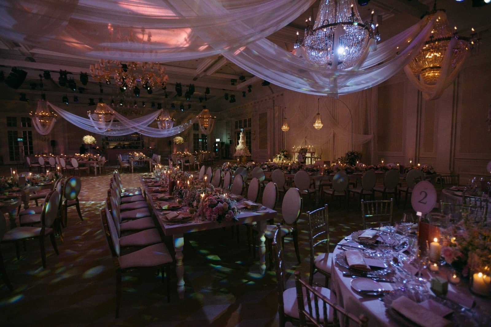 Long dining tables beneath white ceiling drapes and crystal chandeliers, with patterned lighting across the ballroom floor at Raffles London.