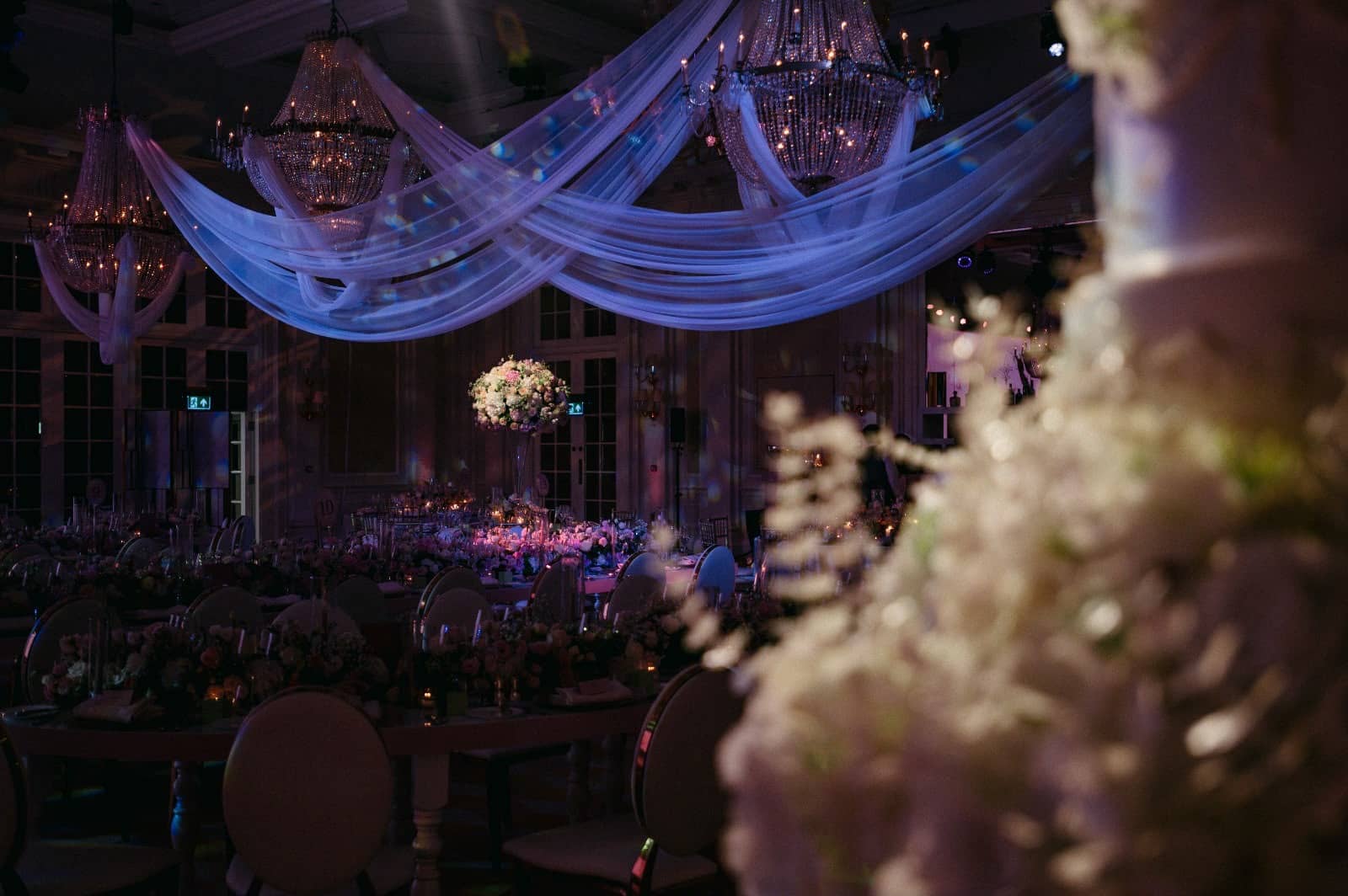 Crystal chandeliers and white ceiling drapes lit in blue above the ballroom, with flowers in the foreground at Raffles London.