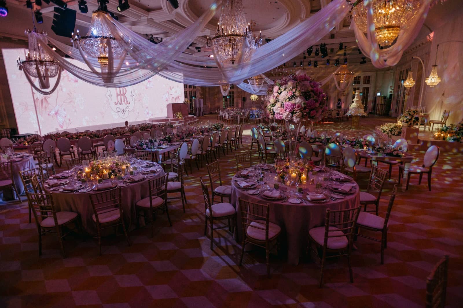 Round dining tables and floral centrepieces beneath chandeliers, with a pink floral LED backdrop across the ballroom at Raffles London.