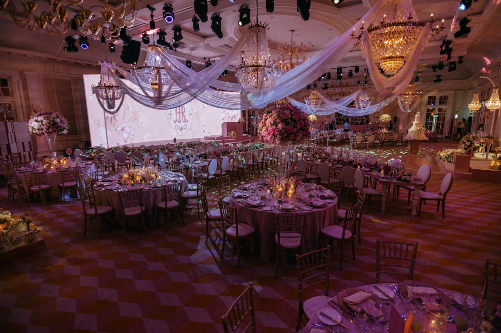 Lighting fixtures and draped chandeliers above a ballroom set with round tables and a wide pink LED screen at Raffles London.