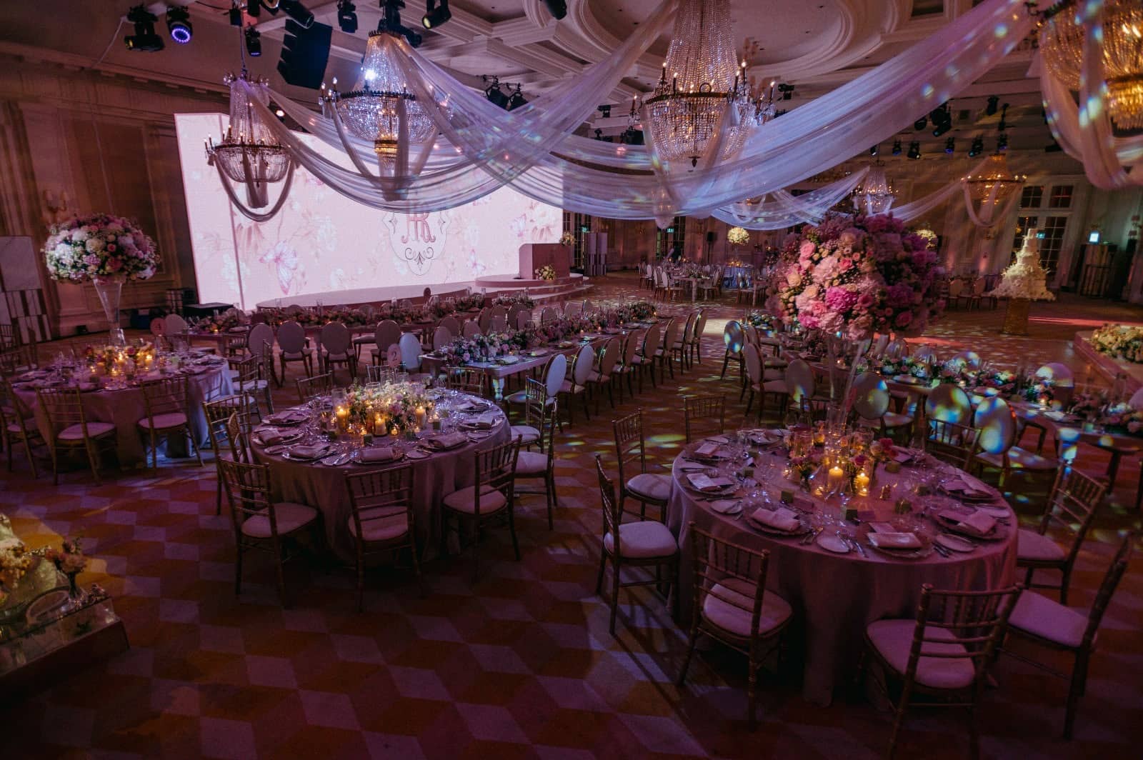 Floral centrepieces rise above ballroom dining tables in front of a wide pink LED screen, beneath white ceiling drapes at Raffles London.