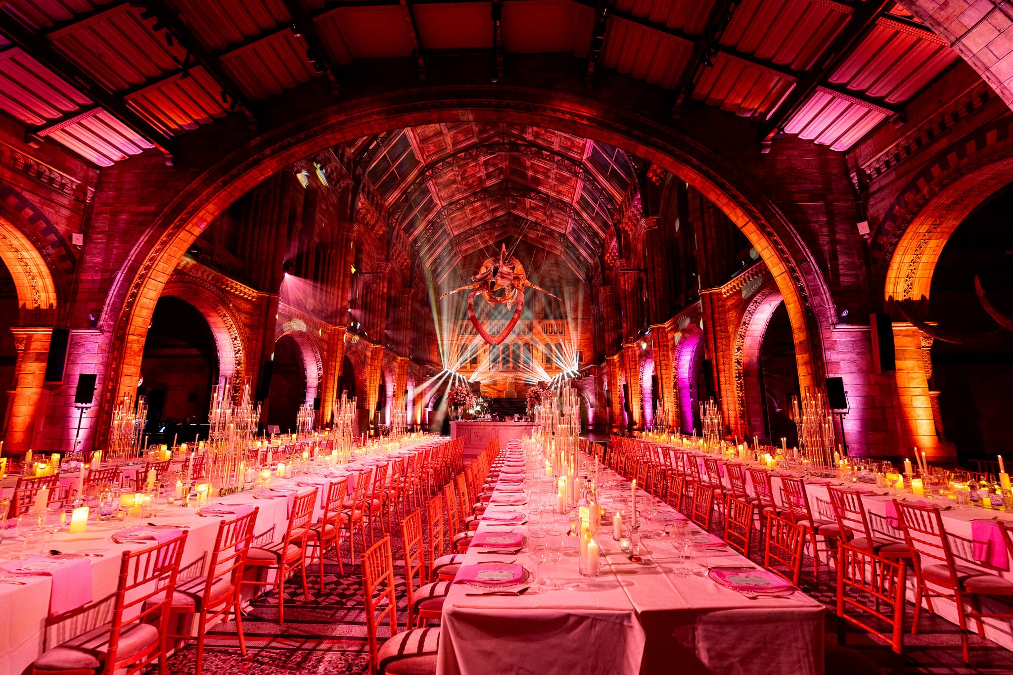 Red uplighting over long tables dressed in pink linen and candles in Hintze Hall, a Natural History Museum wedding dinner lit by Kudos AV.