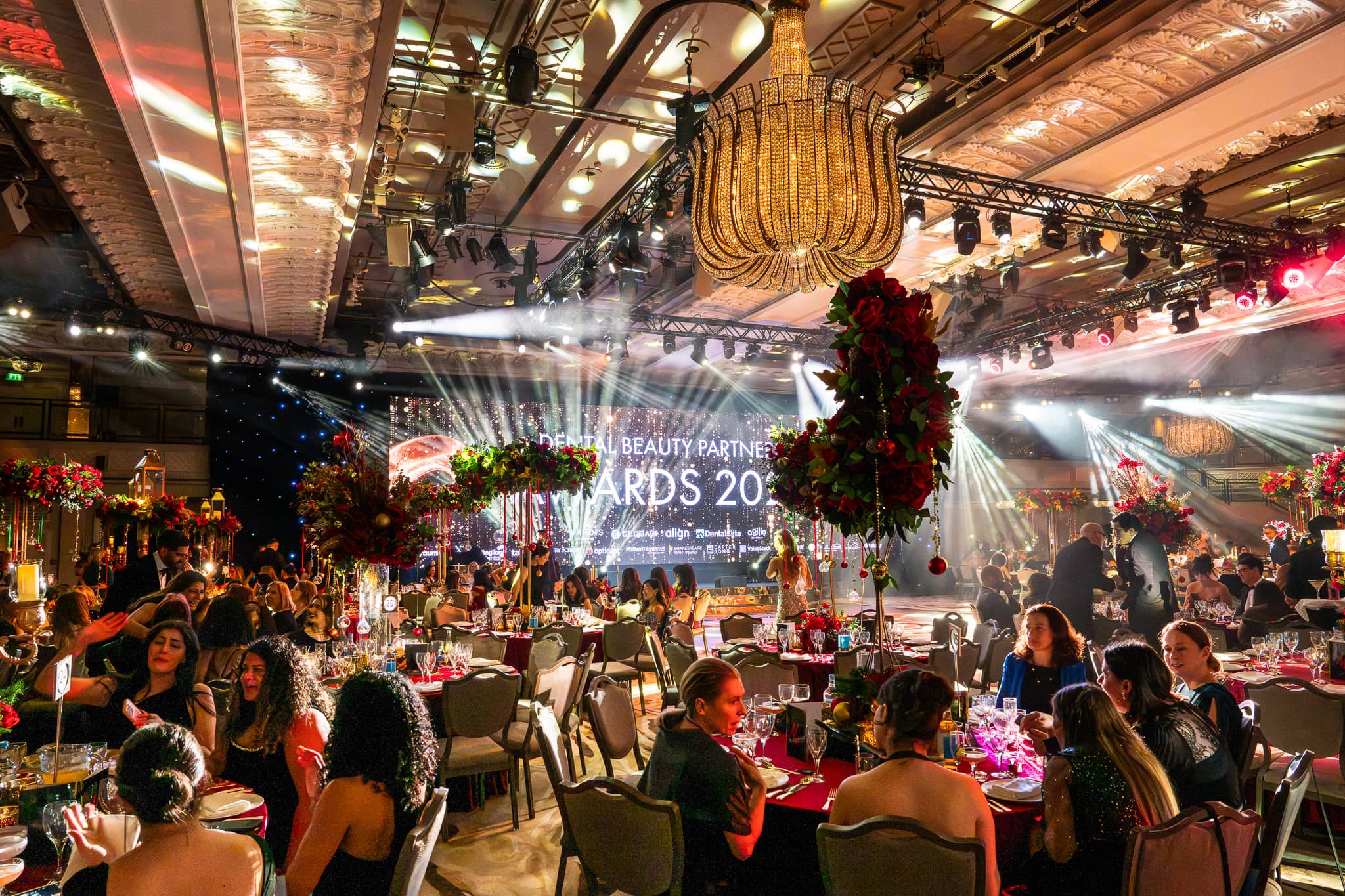 Floral centrepieces in the foreground and the Dental Beauty Partners Awards on the LED screen beyond, dinner lit by Kudos AV in the Great Room at Grosvenor House London.