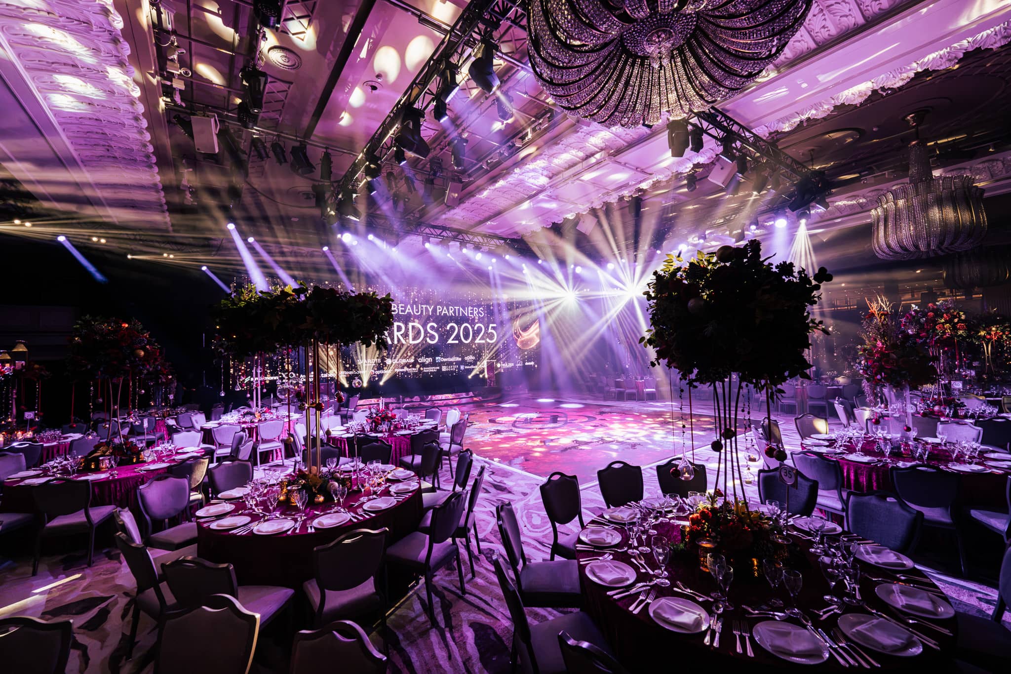 Round tables and floral centrepieces under purple light, the stage screen behind, the Dental Beauty Partners Awards 2025 dinner in the Great Room at Grosvenor House London, by Kudos AV.