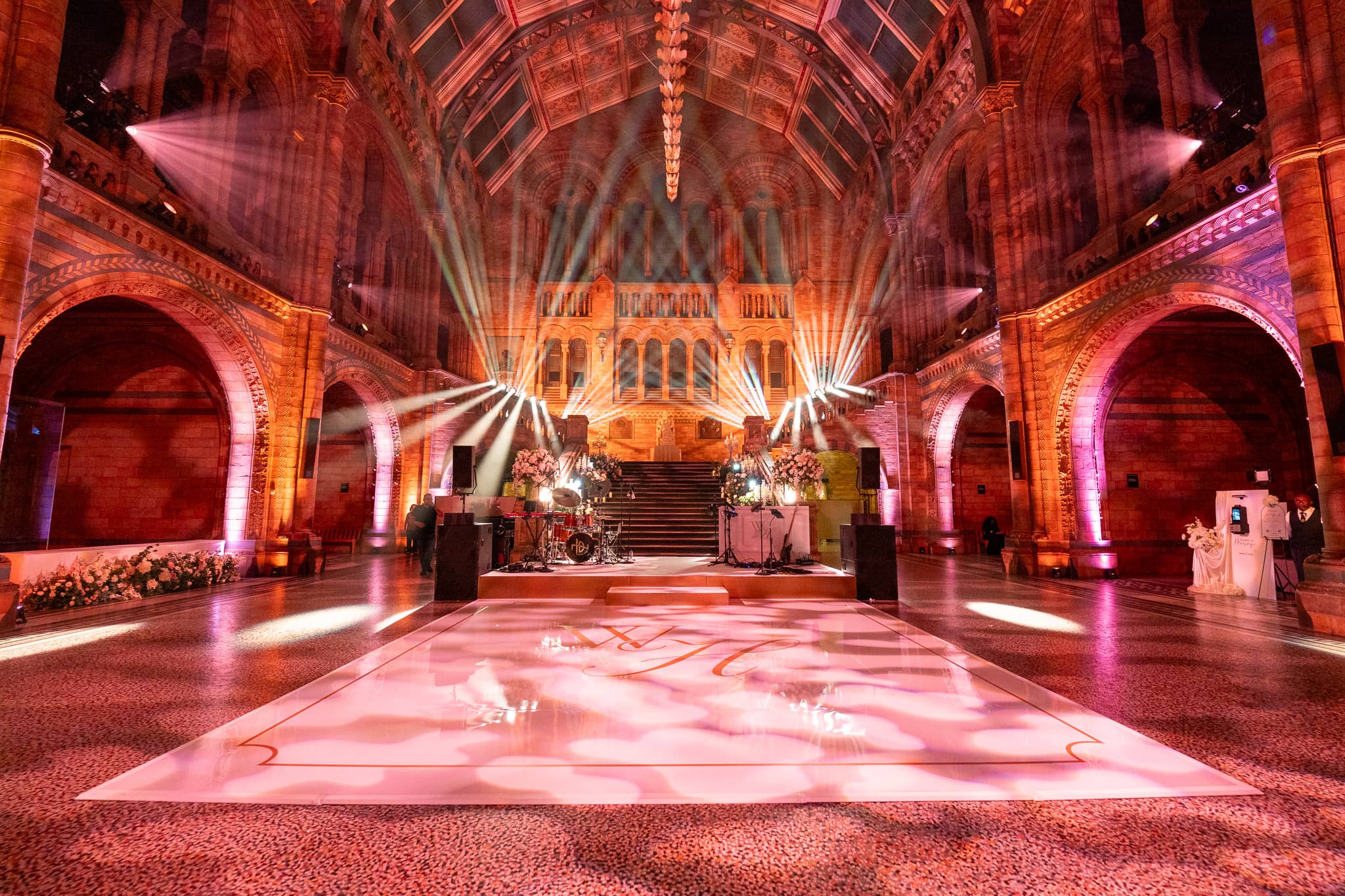 Red and pink uplighting up the arches of Hintze Hall to the vault, the centre of the hall set for the party, at a Natural History Museum wedding by Kudos AV.