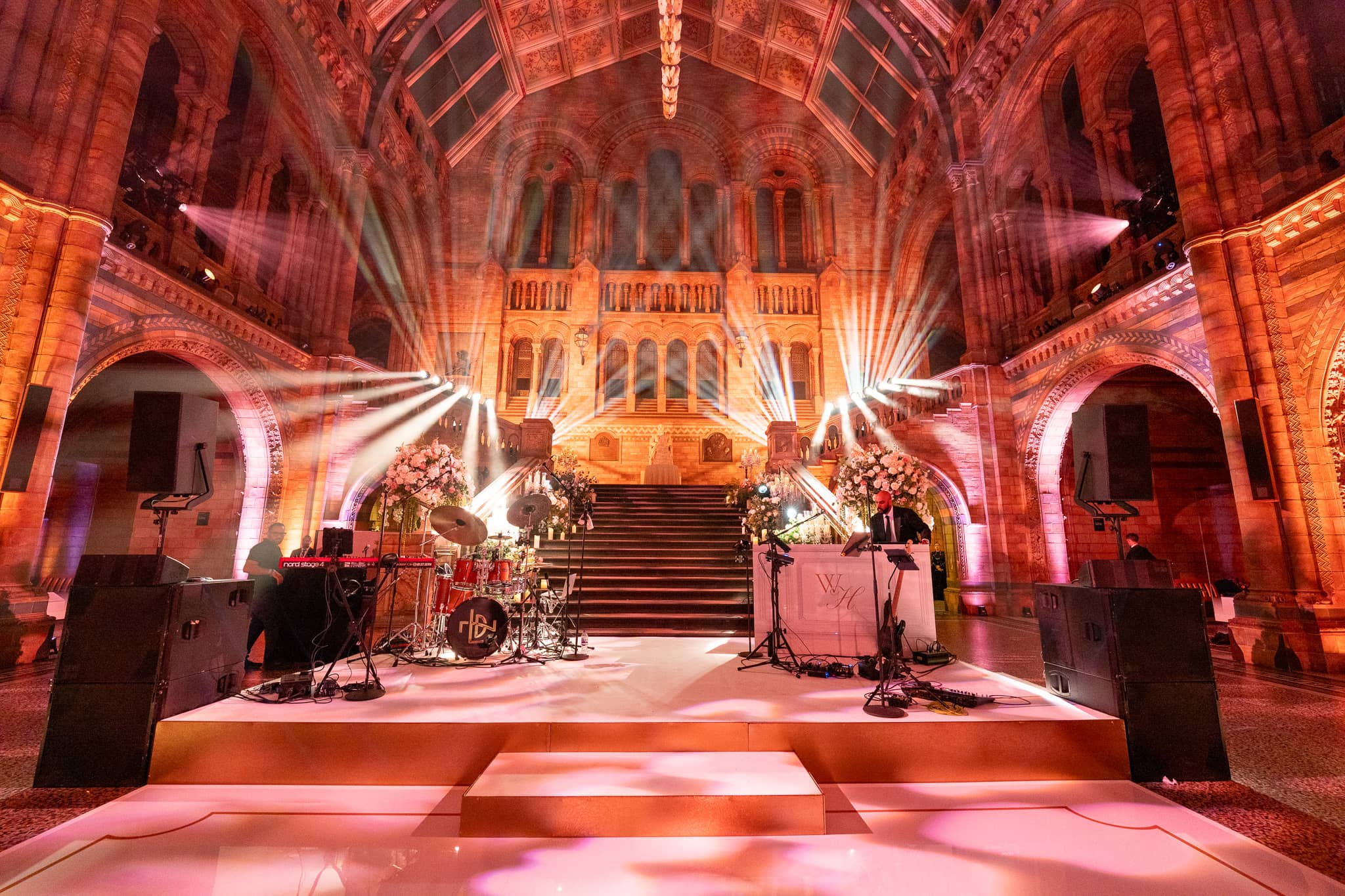 Light beams cutting through the pink and amber washed arches of Hintze Hall at a Natural History Museum wedding, lit by Kudos AV.