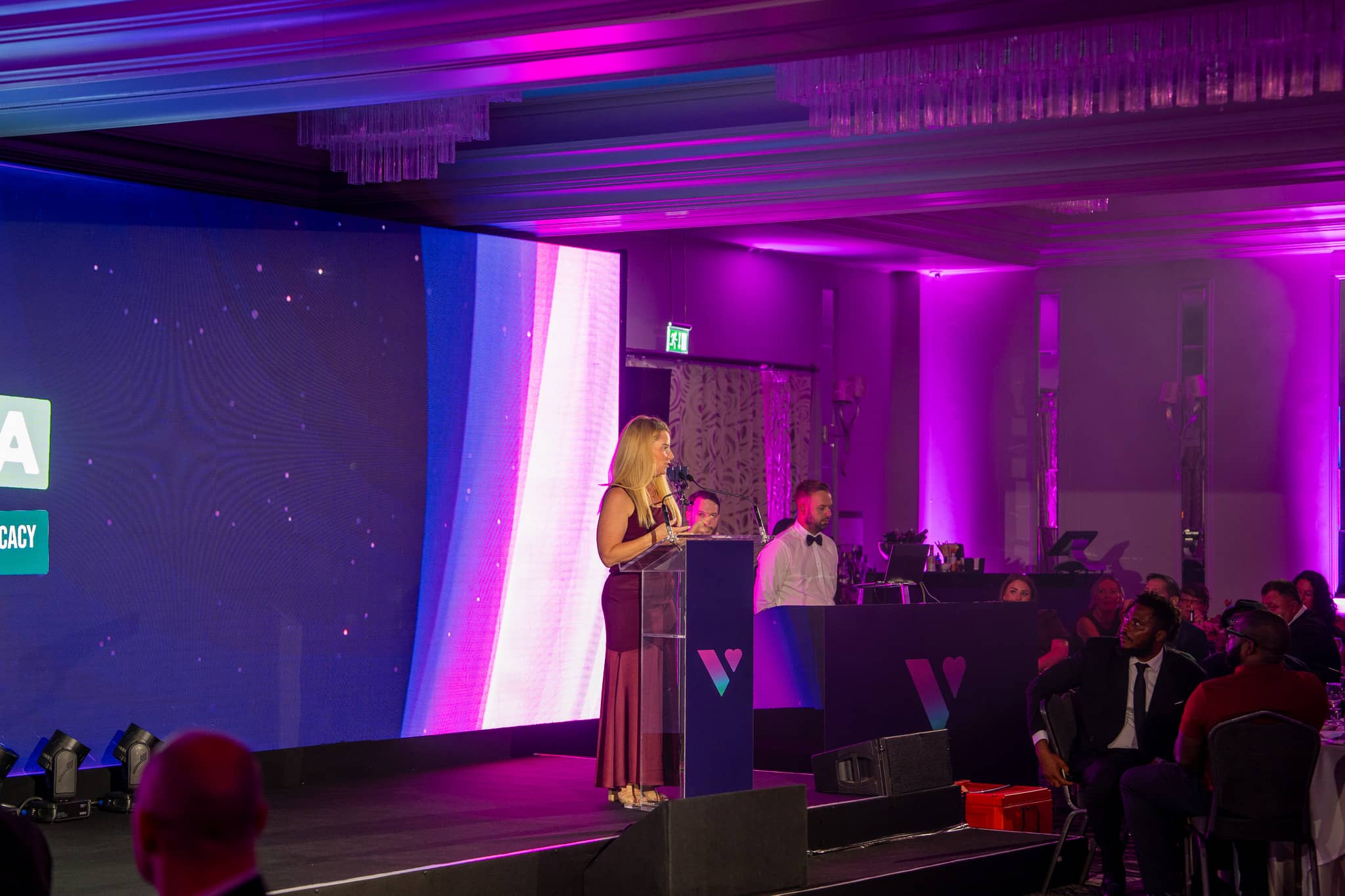 Wider view of a charity presentation at Verve Awards Night, with the speaker at the lectern, the LED wall and pink uplighting in the St Alban Suite.
