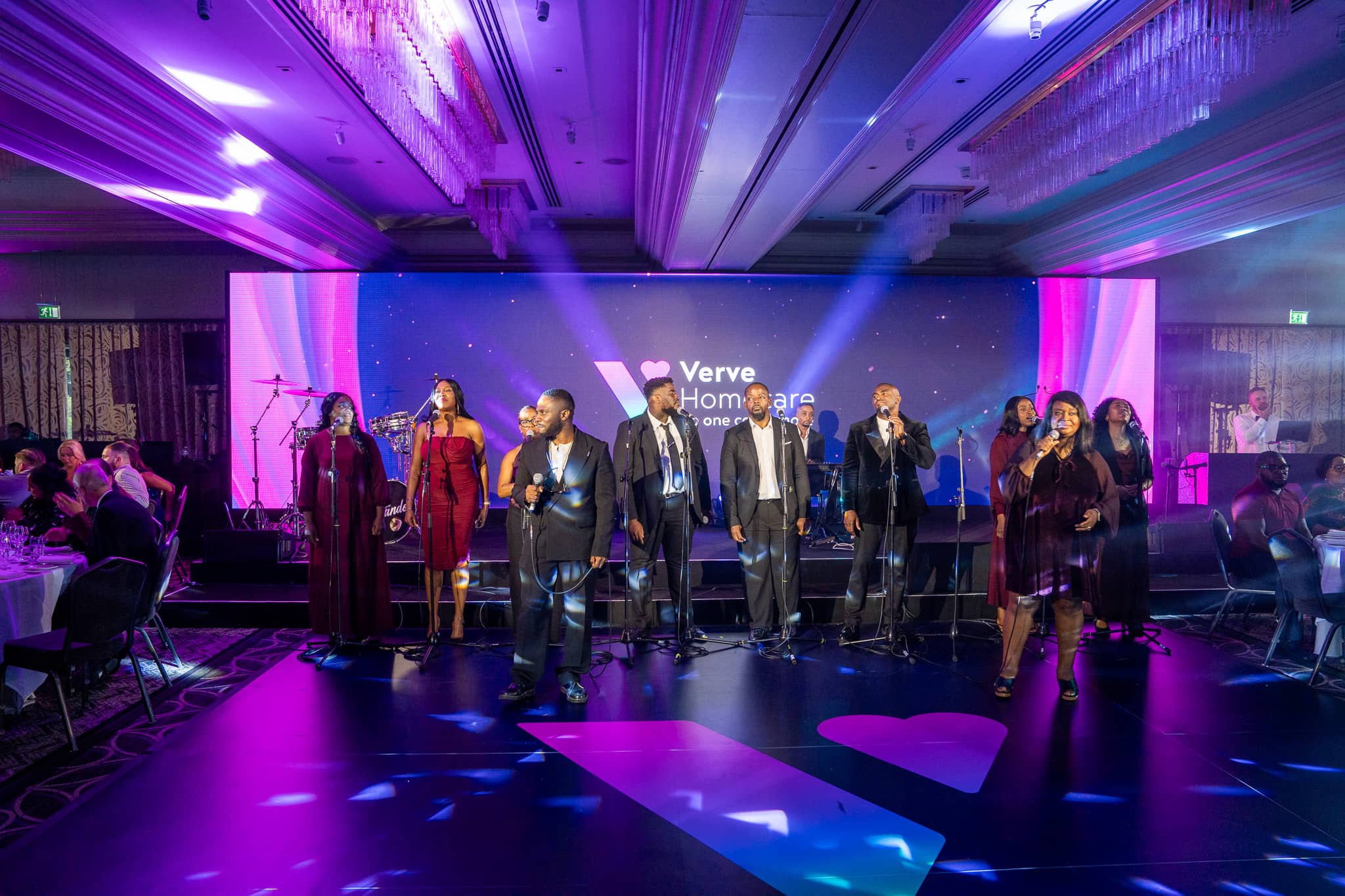 A gospel choir performs on the Verve Awards Night stage at Sopwell House beneath the chandeliers, lit by moving head beams with the Verve logo on the LED wall behind.