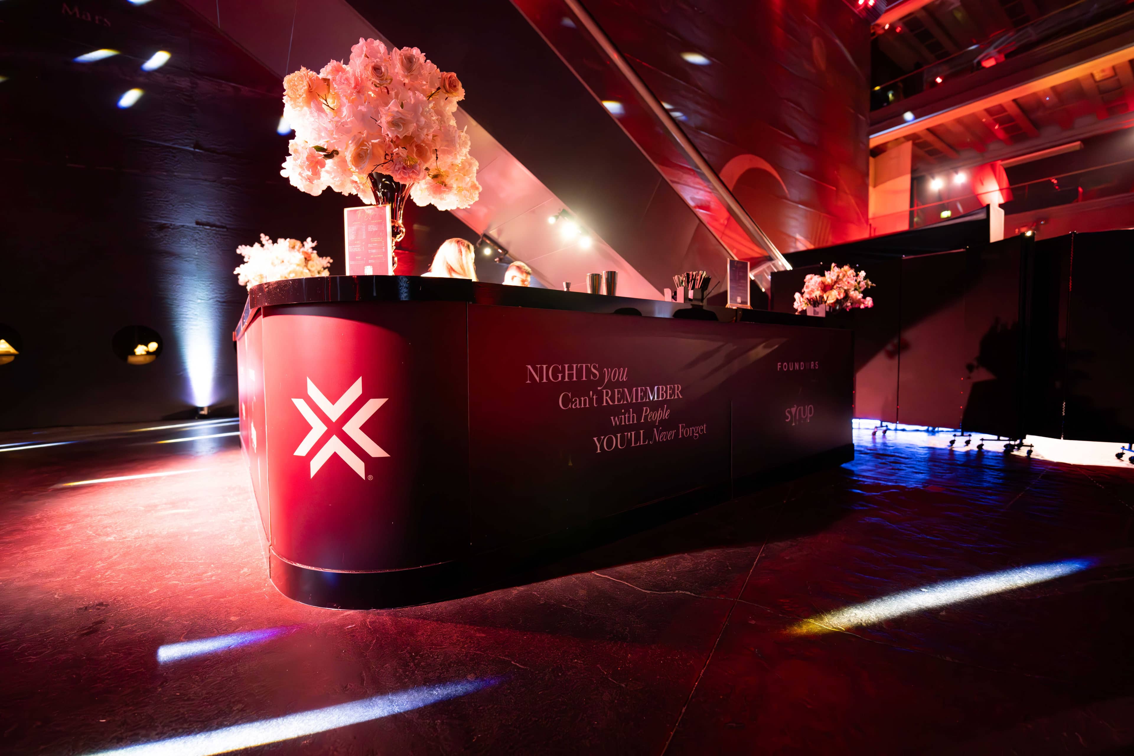 The curved red bar carrying the FOUNDXRS X, pink florals on top and uplighting behind, FOUNDXRS Club black tie party in Earth Hall by Kudos AV.