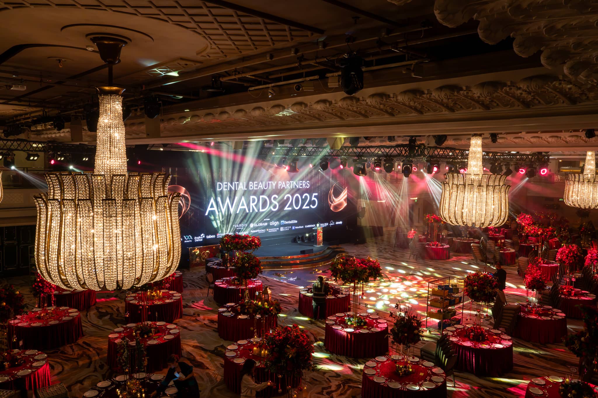 The Great Room at Grosvenor House set for the Dental Beauty Partners Awards 2025: chandeliers, red floral centrepieces, the LED screen on stage and the room lit by Kudos AV.