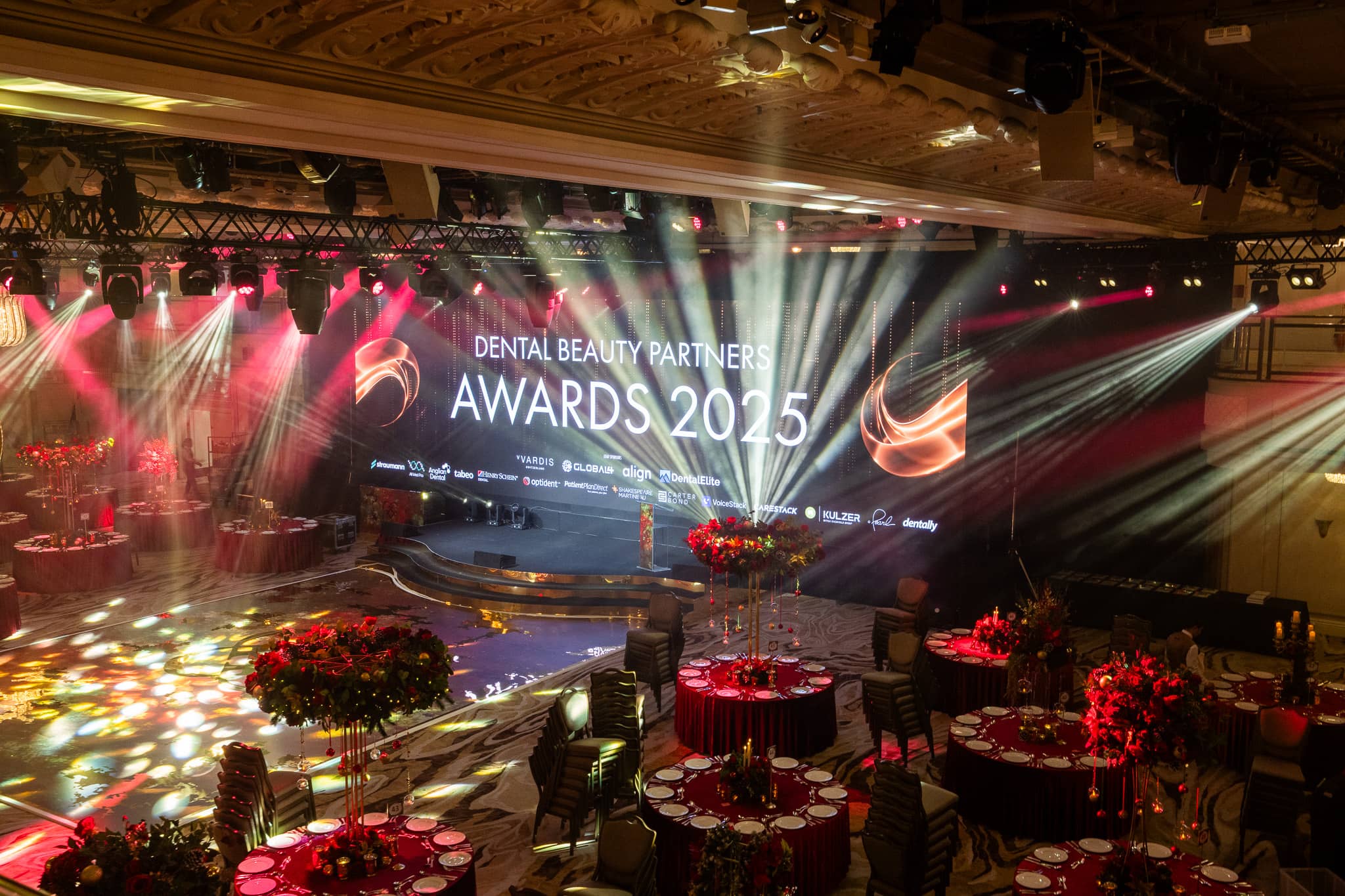 Pink stage lighting and the LED screen over the awards stage, red florals and dressed tables in front, Dental Beauty Partners Awards 2025 in the Great Room at Grosvenor House London, by Kudos AV.
