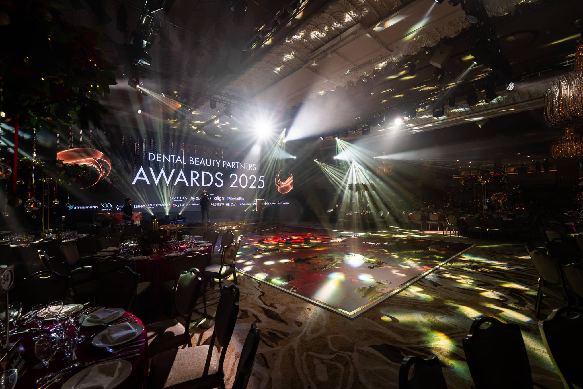 The awards stage and LED screen seen from the dinner tables, beams cutting across the room, Dental Beauty Partners Awards 2025 in the Great Room at Grosvenor House London, by Kudos AV.