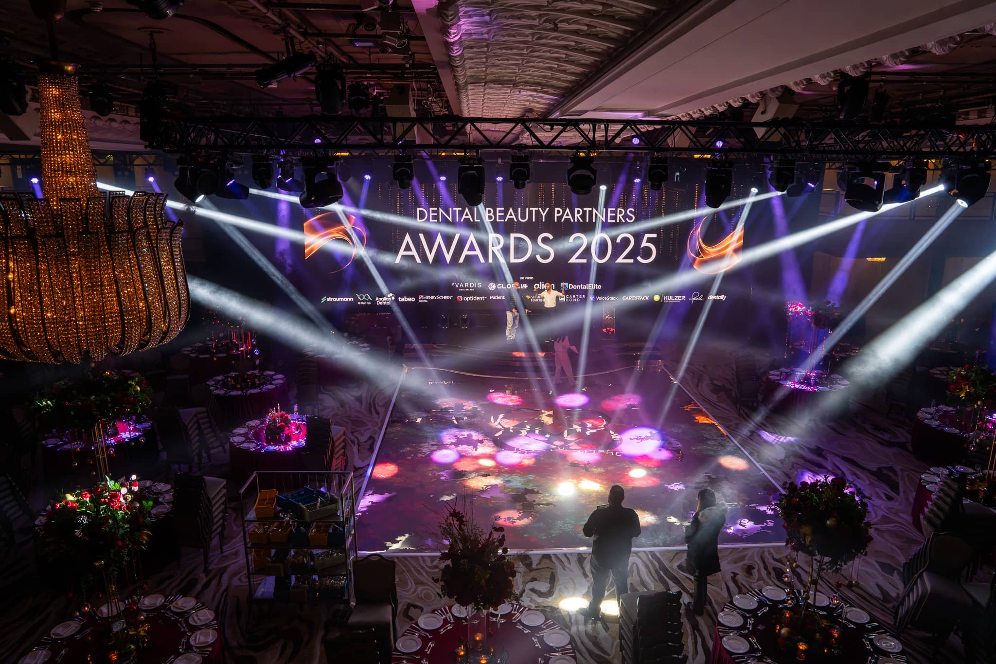 Purple and white stage lighting around the LED screen showing the Dental Beauty Partners Awards 2025 title, with red florals on the tables, in the Great Room at Grosvenor House London, by Kudos AV.