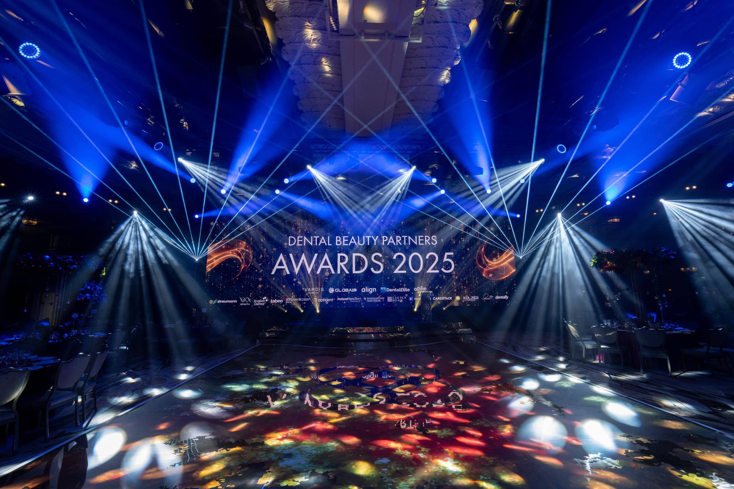 Blue uplighting around the Great Room, white beams over the stage and the printed dancefloor lit in front, Dental Beauty Partners Awards 2025 at Grosvenor House London by Kudos AV.