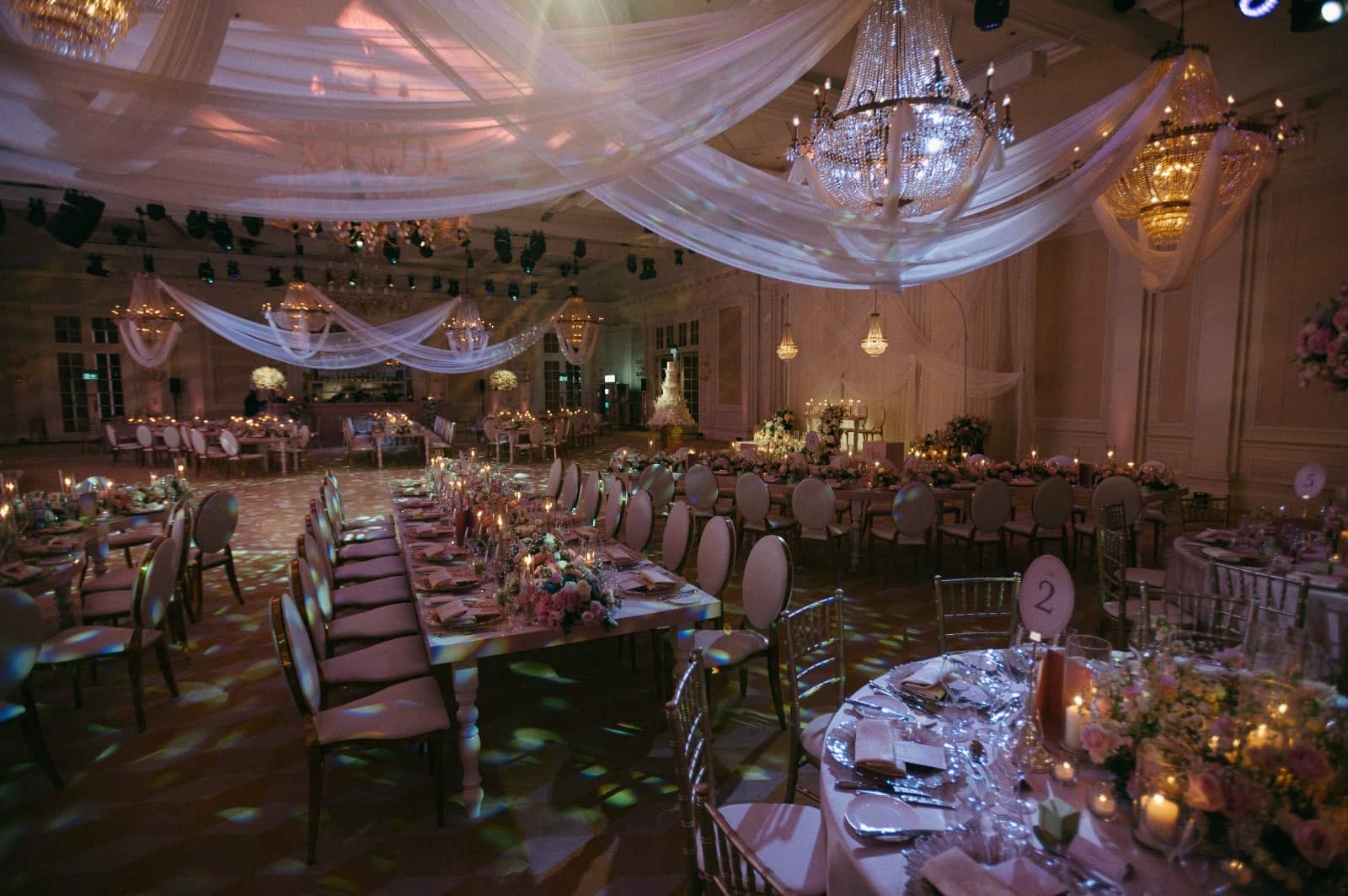 A candlelit table setting beside rows of ballroom dining tables, with chandeliers and patterned lighting on the floor at Raffles London.