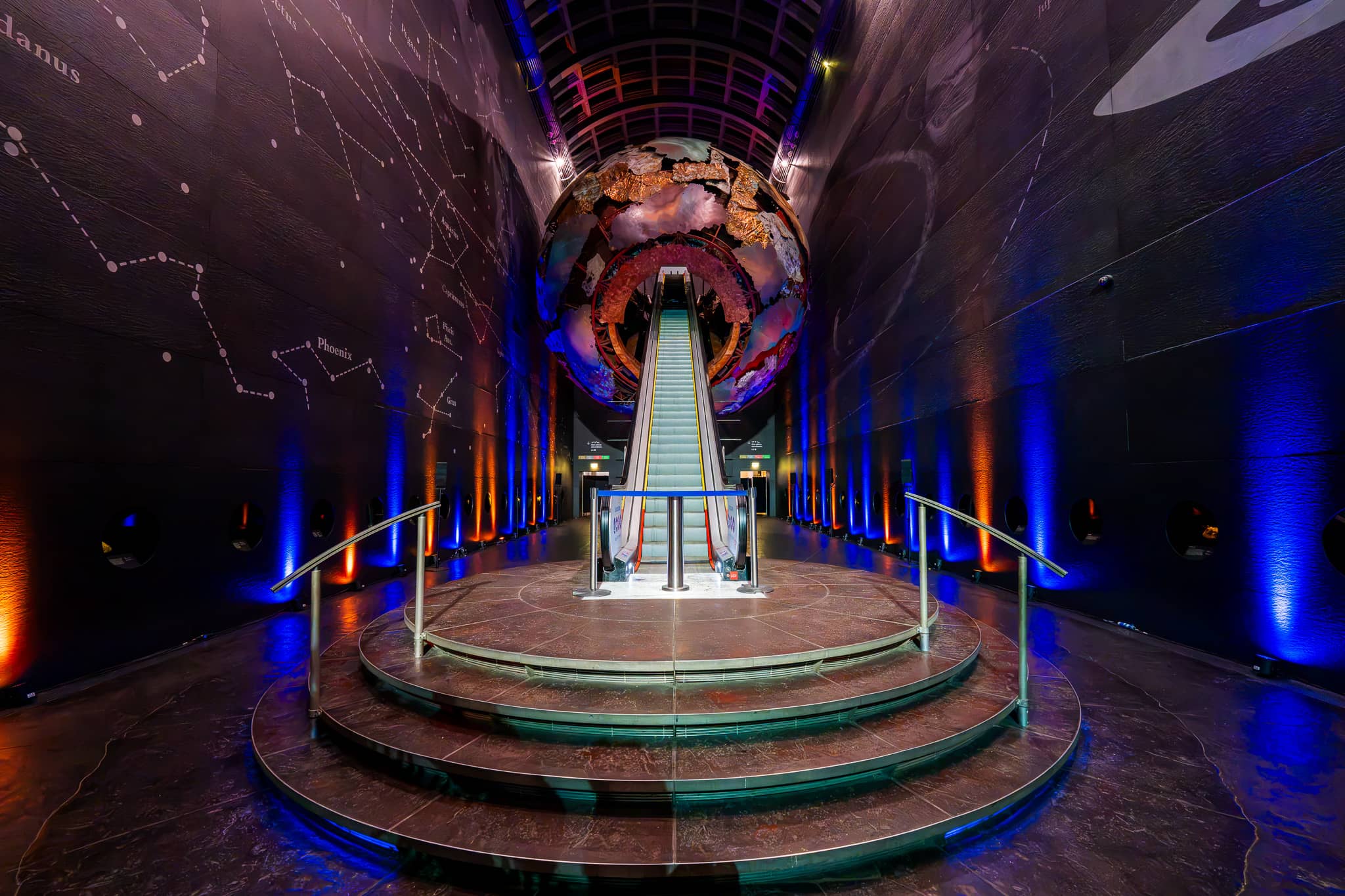 The escalator in Earth Hall rising into the Earth sculpture, the walls lit blue and amber for the Yes Theory 10 million subscriber celebration at the Natural History Museum, by Kudos AV.