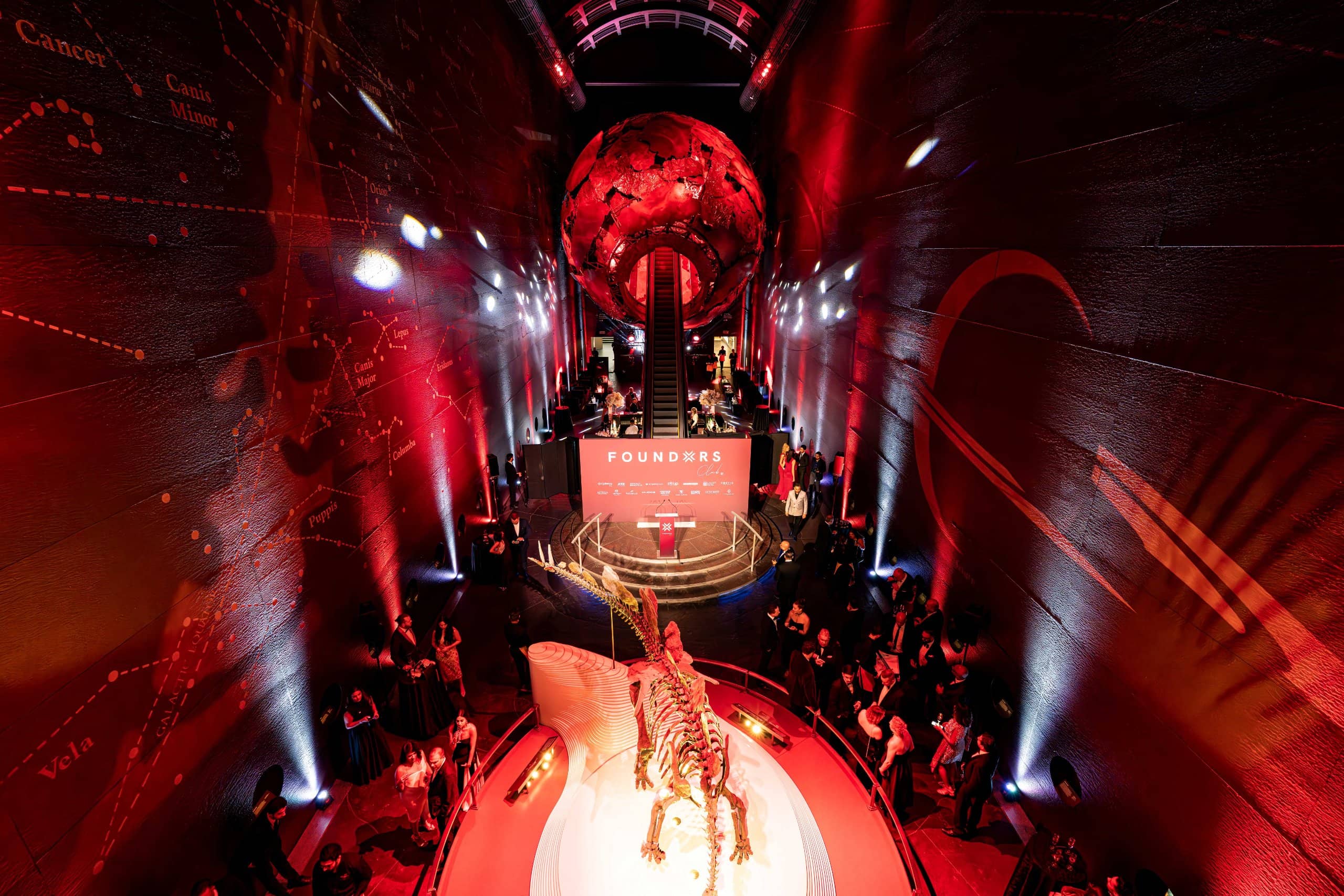 Earth Hall washed red with guests around the stage and the Earth sculpture lit above, FOUNDXRS Club black tie party at the Natural History Museum, by Kudos AV.
