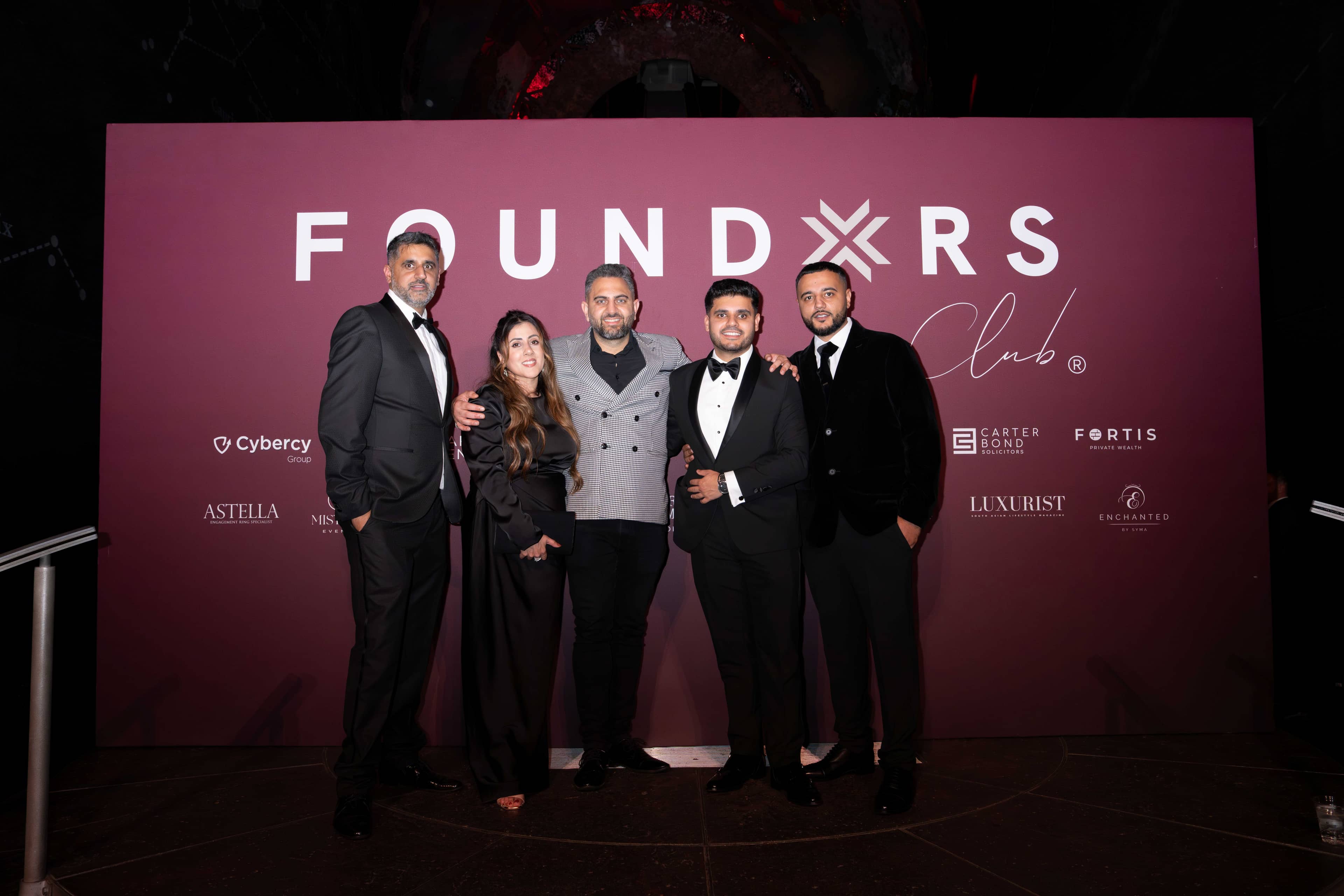 Five guests in black tie at the FOUNDXRS Club backdrop with the evening’s partners, at the FOUNDXRS Club black tie party in Earth Hall, Natural History Museum.