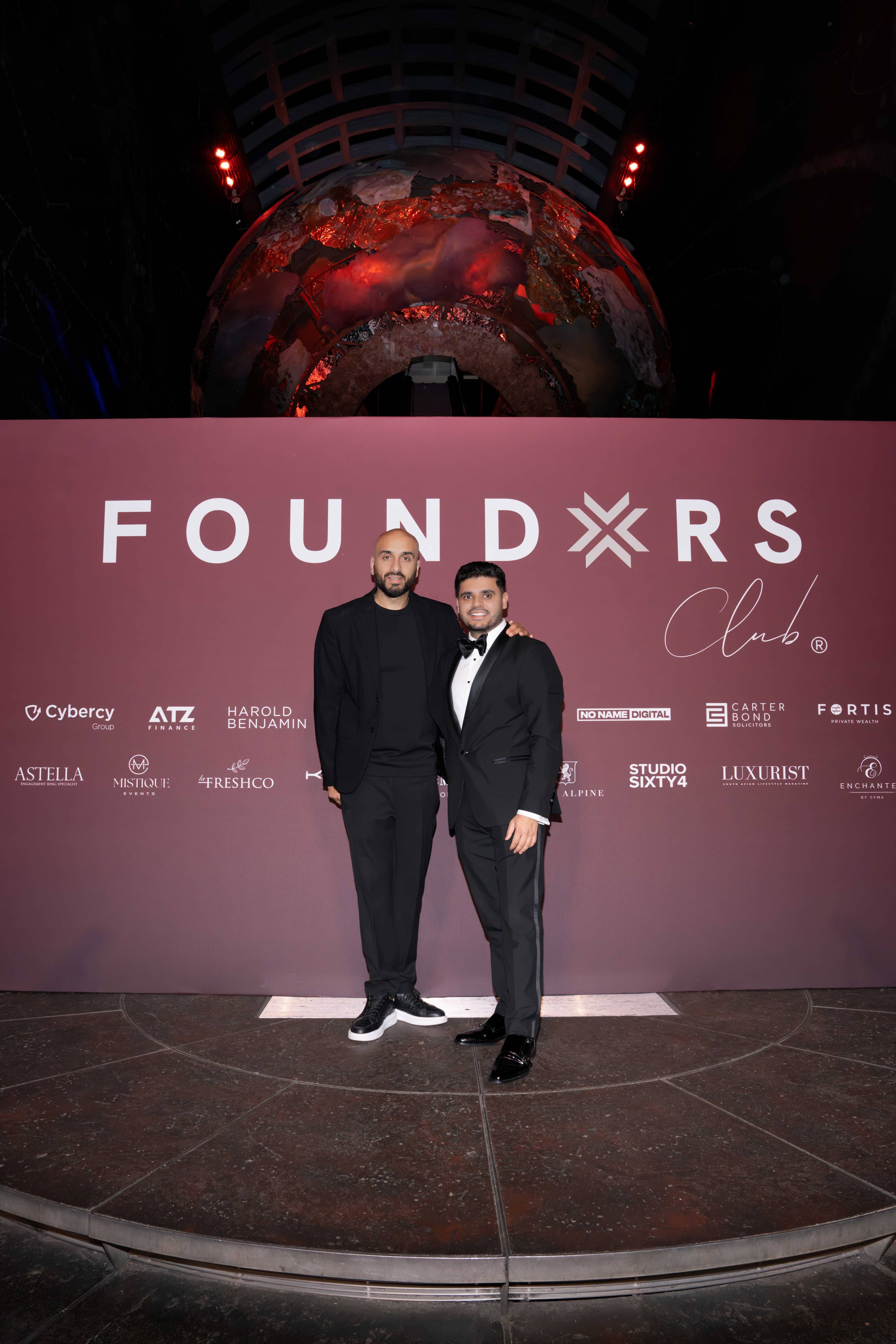 Two guests in black tie at the FOUNDXRS Club backdrop, the Earth sculpture lit above them, FOUNDXRS Club black tie party in Earth Hall, by Kudos AV.