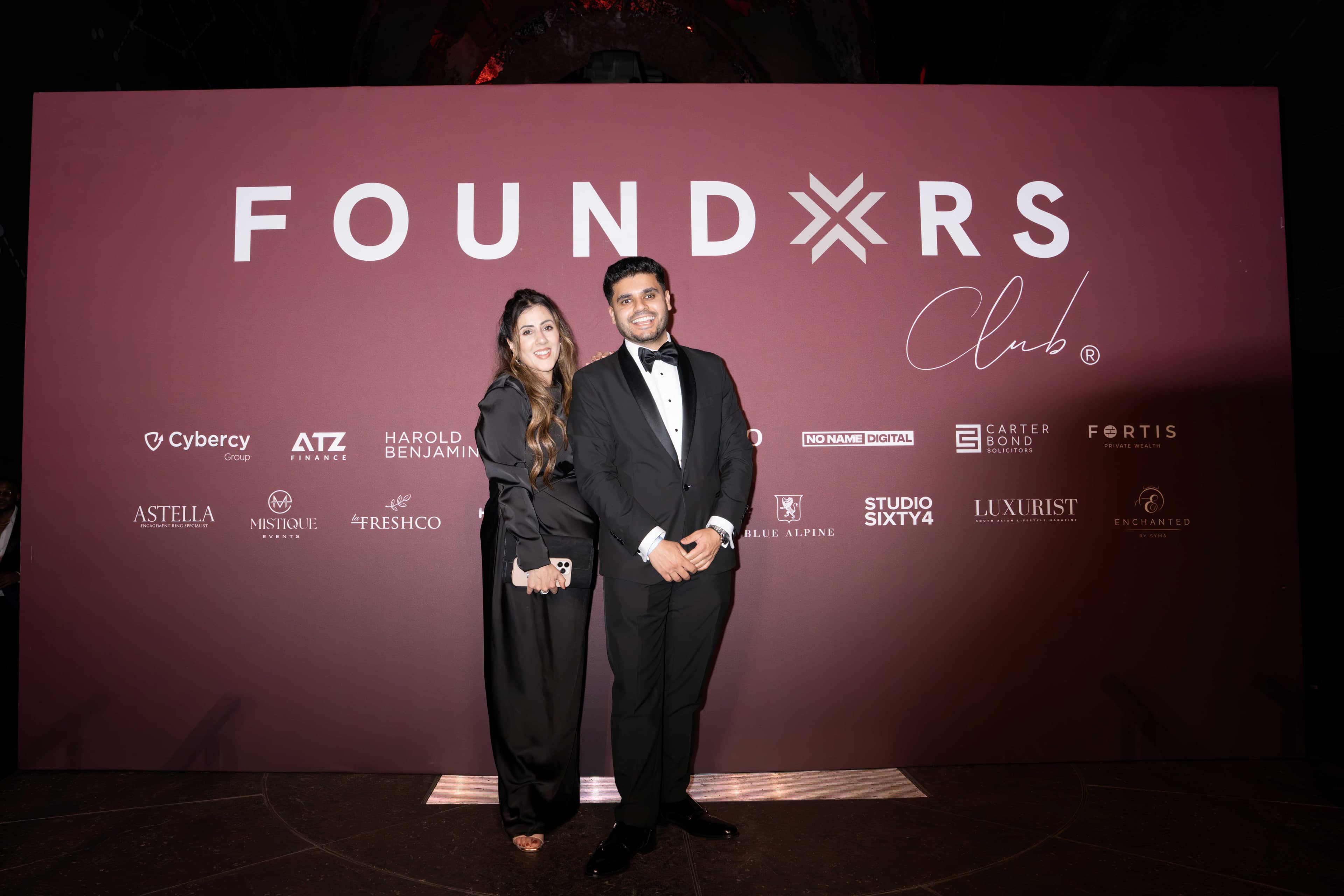 Two guests in black tie in front of the FOUNDXRS Club backdrop at the FOUNDXRS Club black tie party, Earth Hall, Natural History Museum.