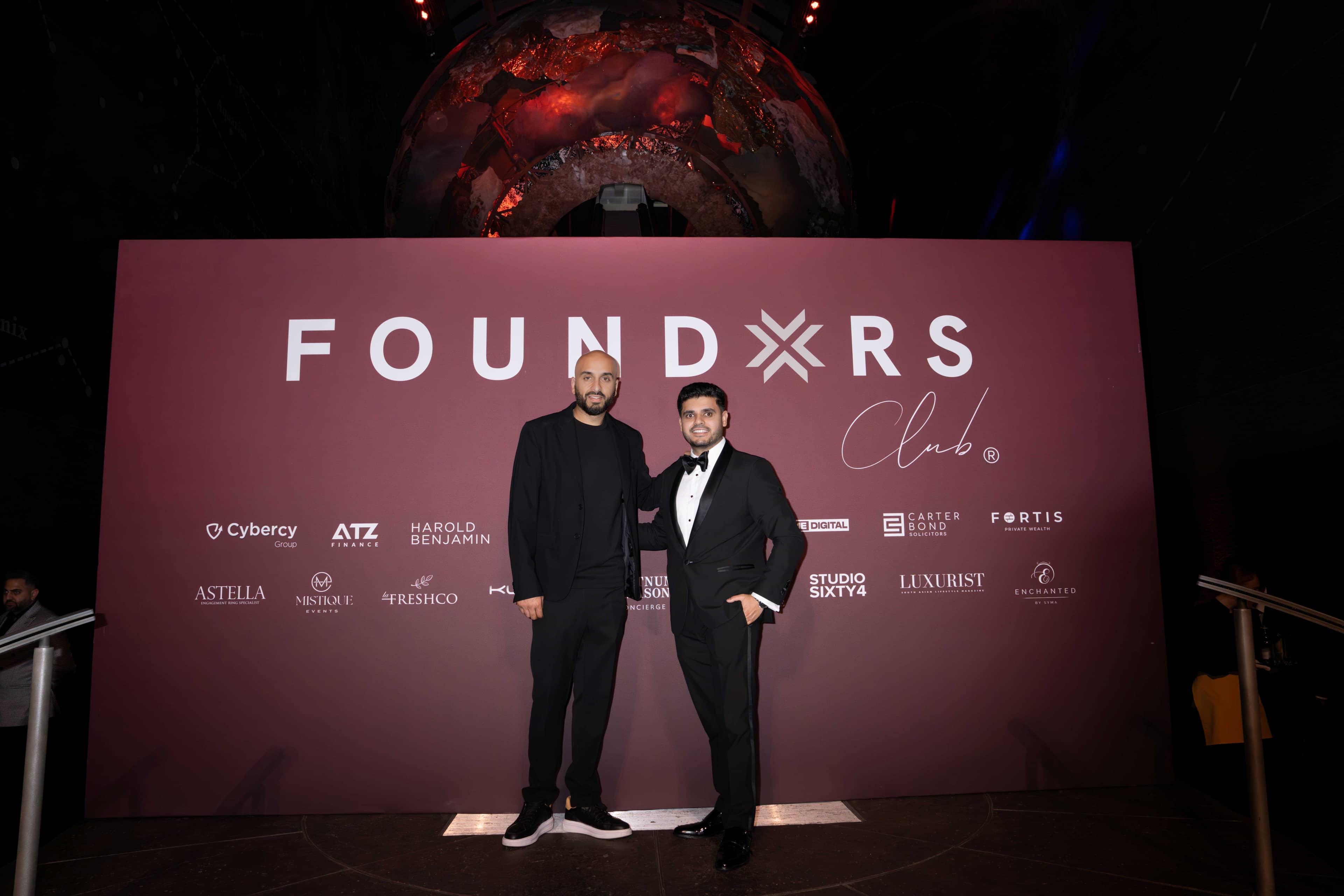 Two guests at the FOUNDXRS Club backdrop with the partner logos, the Earth sculpture glowing above, FOUNDXRS Club black tie party in Earth Hall by Kudos AV.