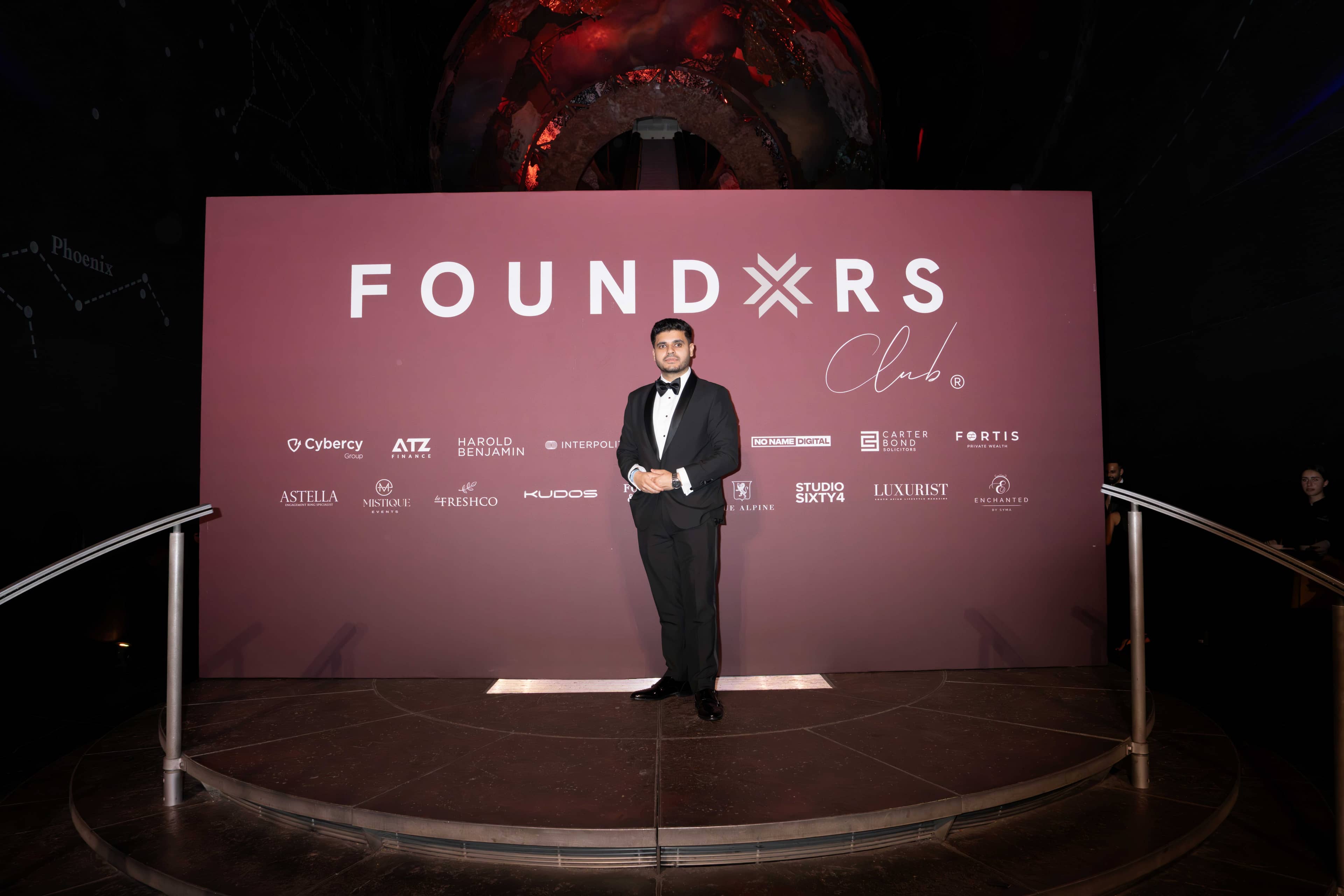 A guest in black tie on stage in front of the screen carrying the FOUNDXRS Club branding and partners, FOUNDXRS Club black tie party in Earth Hall by Kudos AV.