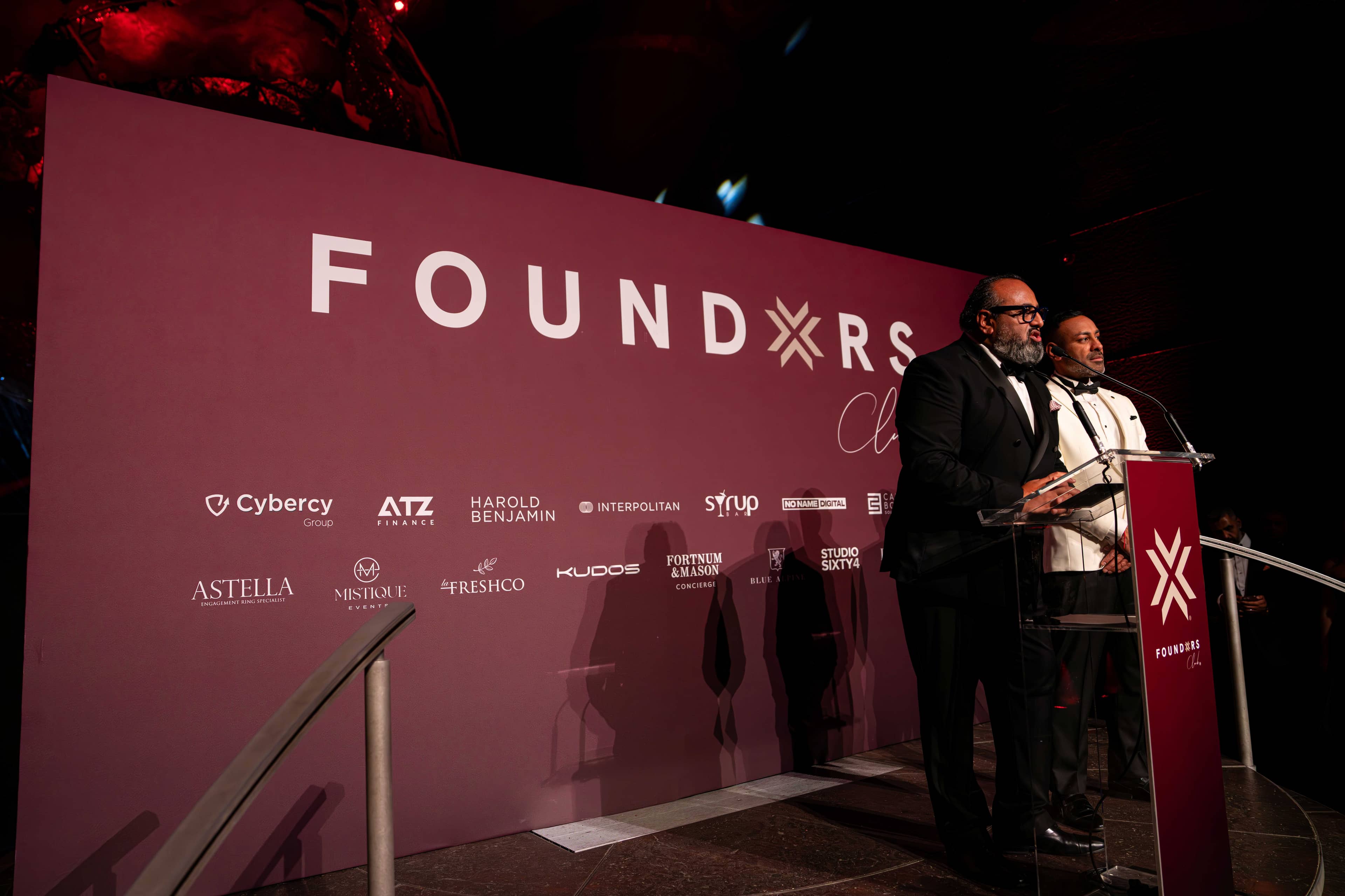 Two speakers at the red lectern in front of the screen carrying FOUNDXRS Club and its partners, FOUNDXRS Club black tie party in Earth Hall by Kudos AV.