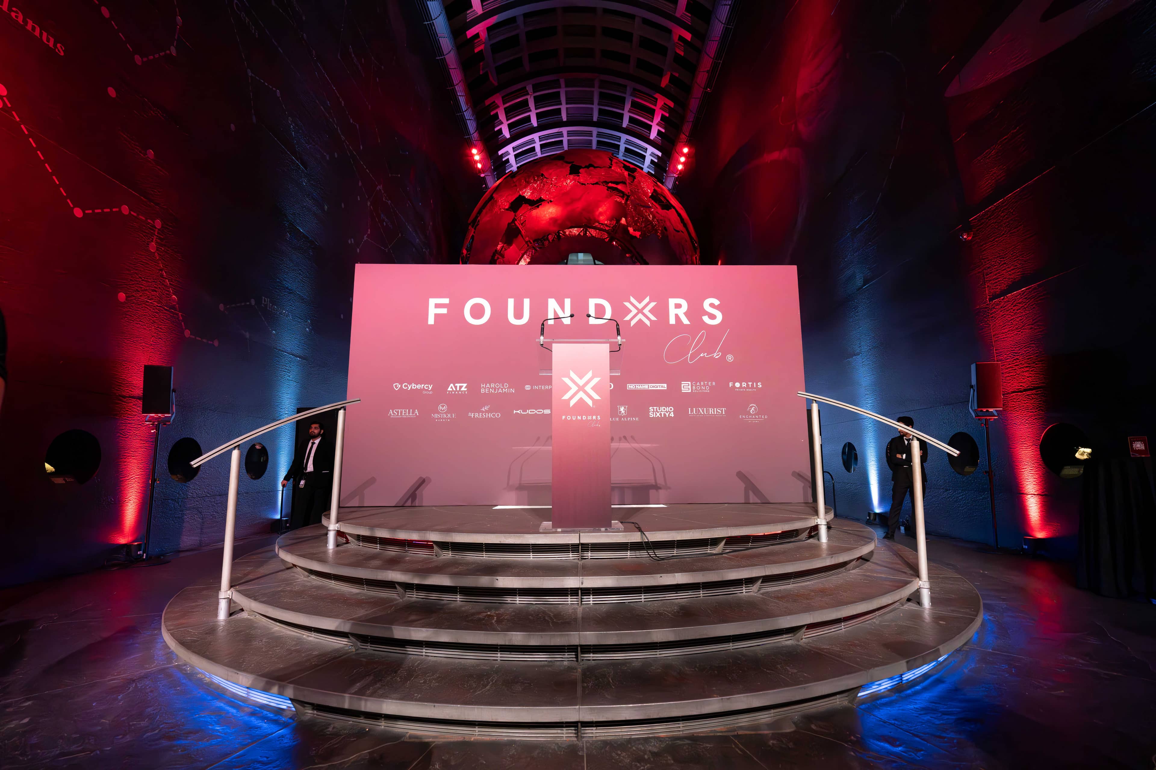 The FOUNDXRS Club stage lit pink, red and blue uplighting and the curved balcony rails of Earth Hall around it, FOUNDXRS Club black tie party by Kudos AV.