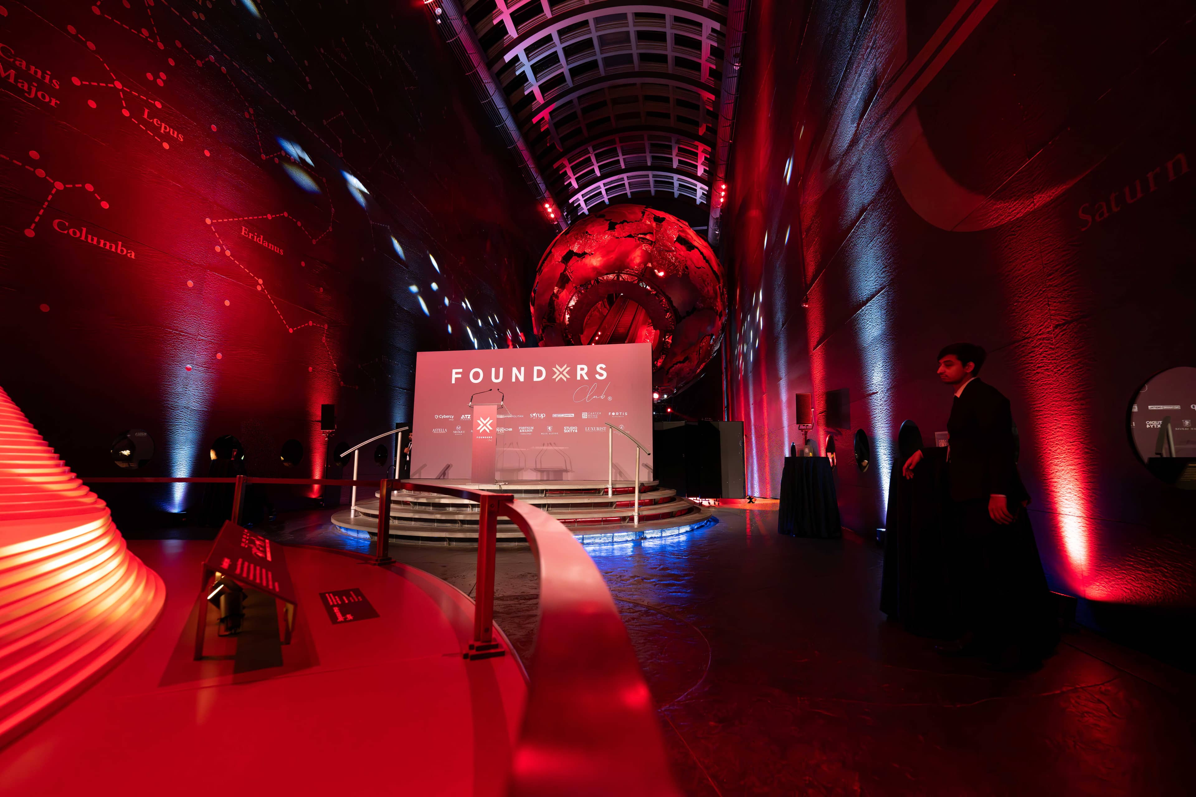 The stage and screen beneath the Earth sculpture in Earth Hall, the walls lit red for the FOUNDXRS Club black tie party, by Kudos AV.