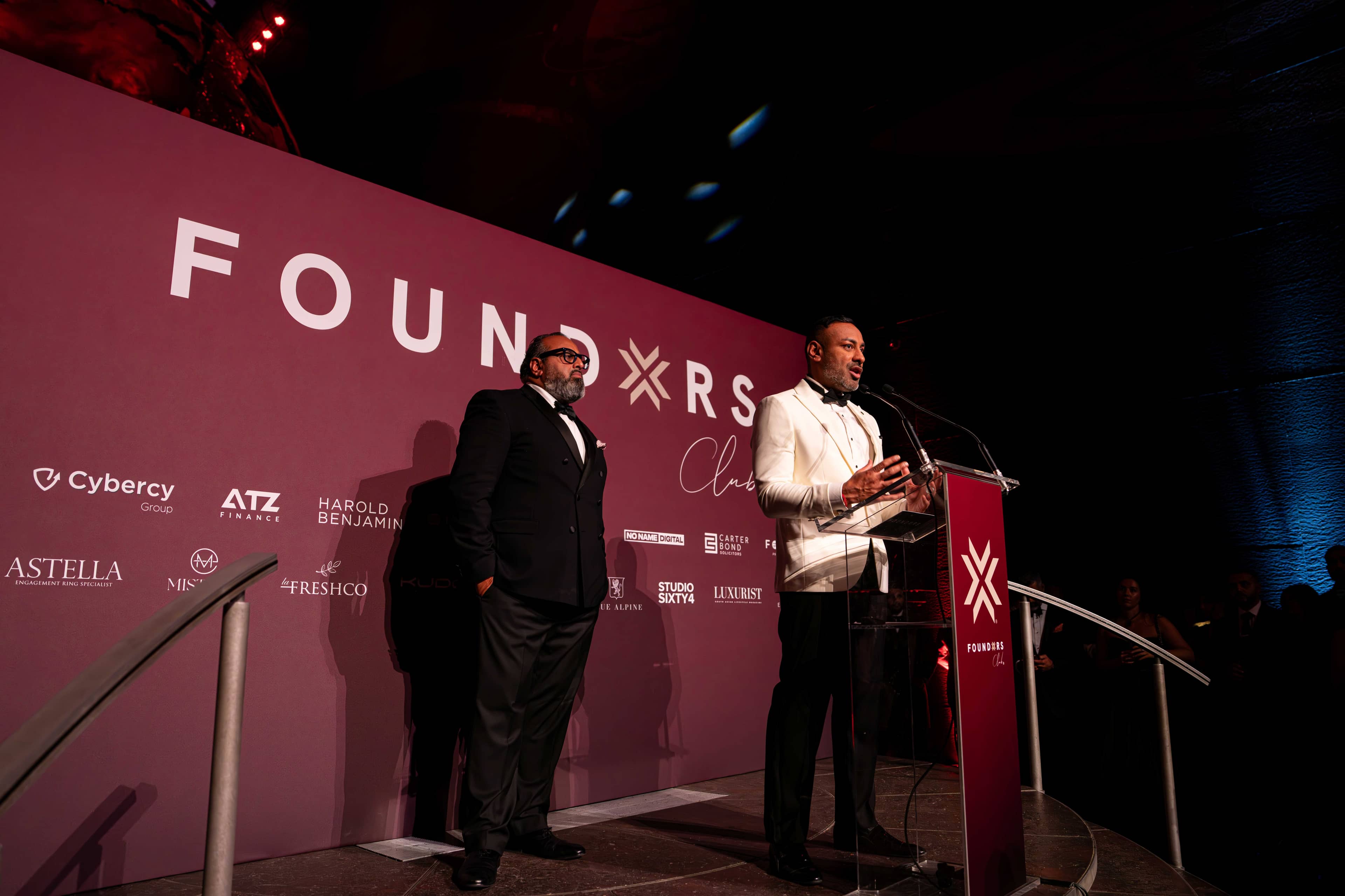 A speaker at the red lectern with a second guest on stage, FOUNDXRS branding behind, FOUNDXRS Club black tie party in Earth Hall by Kudos AV.