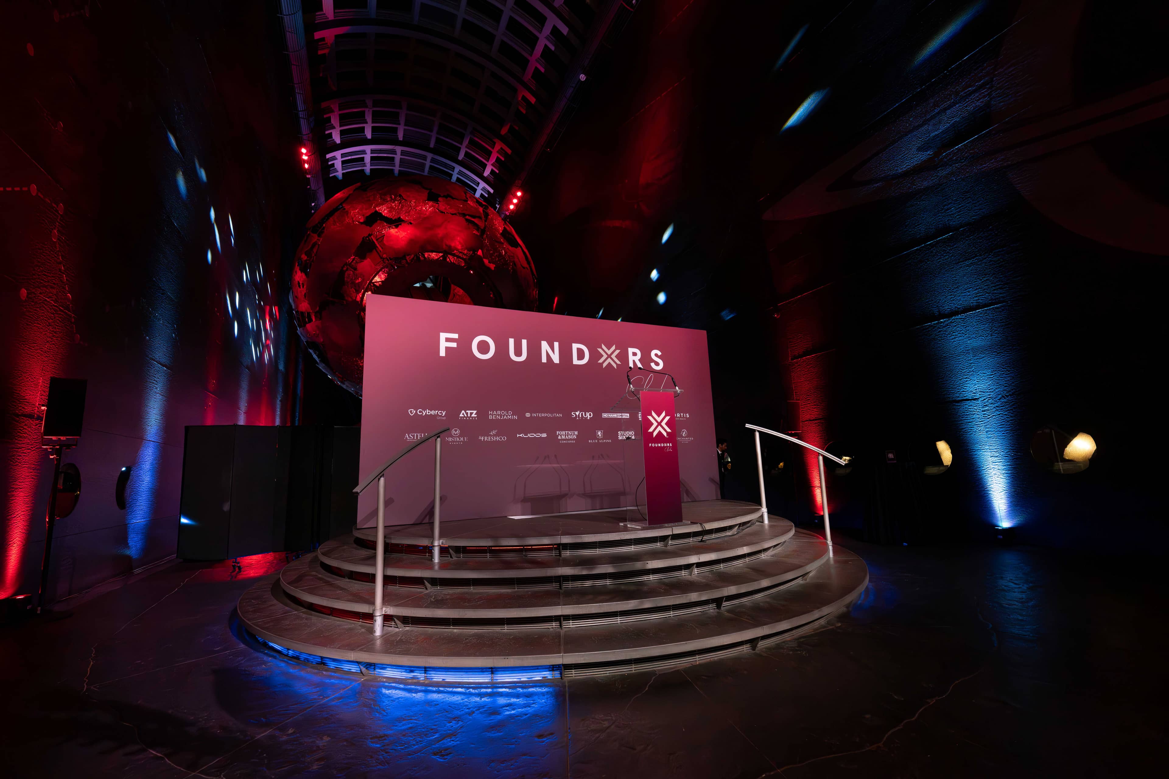 The stage with FOUNDXRS branding in Earth Hall, red uplighting on the walls and blue light along the curved balcony rails, FOUNDXRS Club black tie party by Kudos AV.